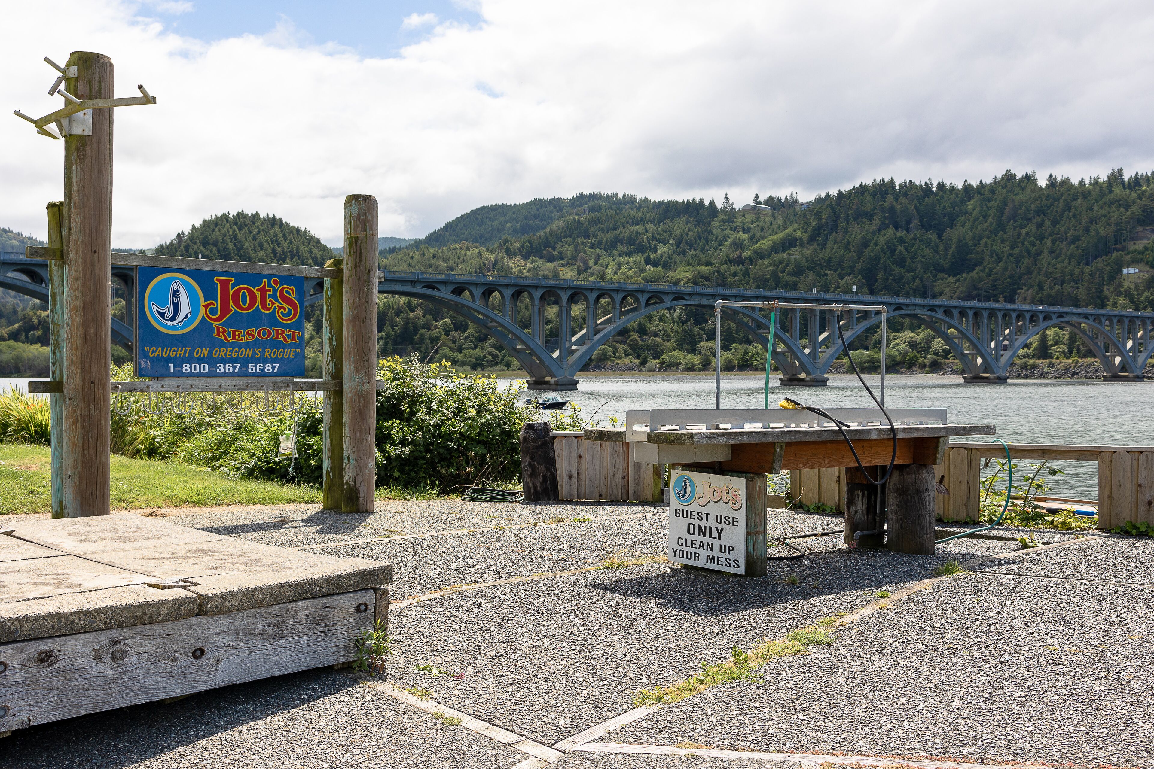 A wooden dock with a sign for Jot's Resort, a bridge in the background, and a forested hillside.