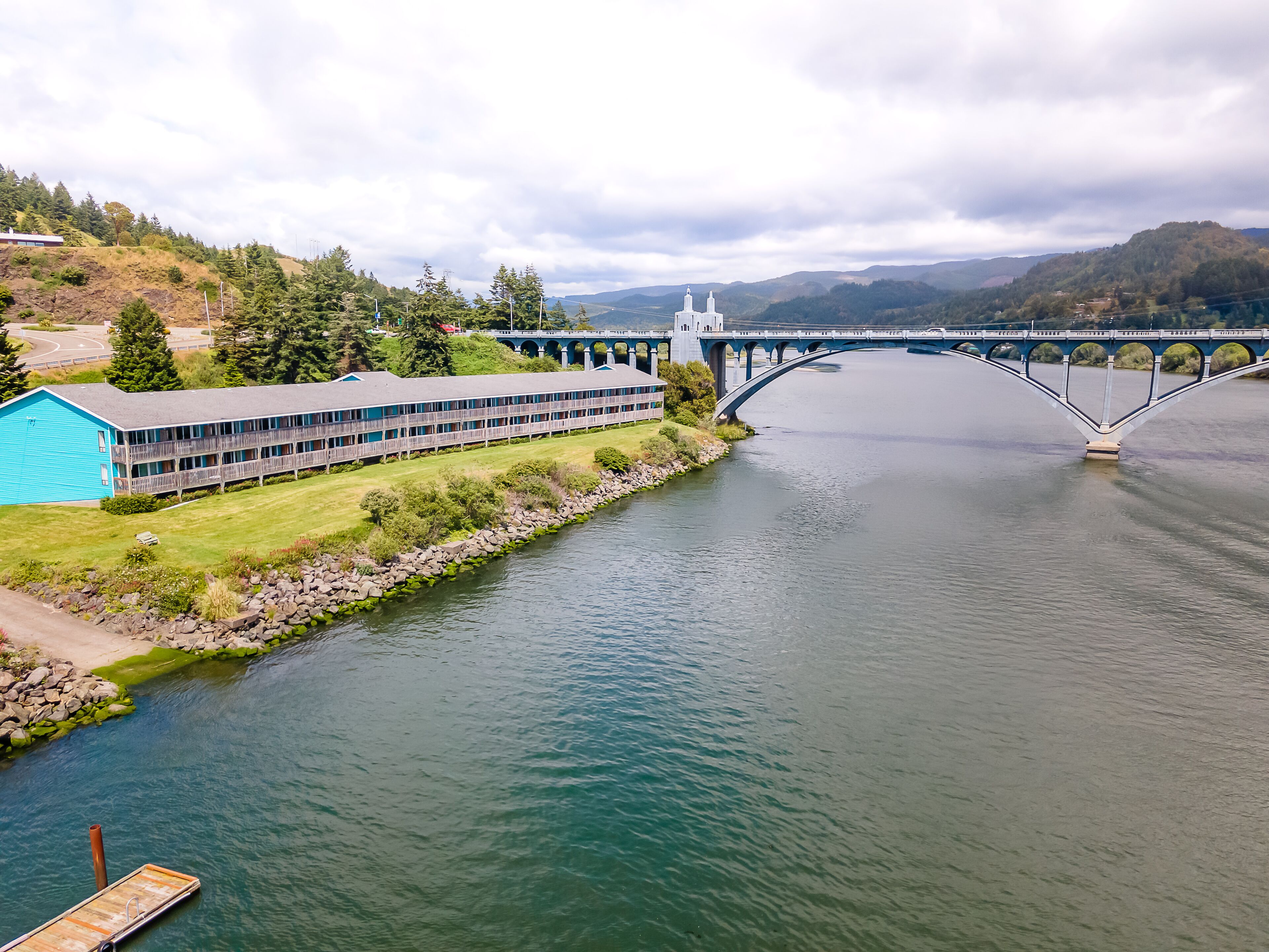 Un grand bâtiment à la structure longue et étroite, doté d’un toit plat, situé à proximité d’une rivière, avec un pont visible au loin.
