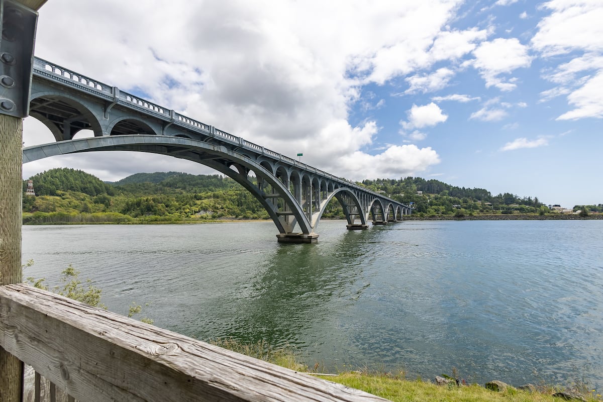 A large, arched bridge over a calm river with a forested hillside in the background.
