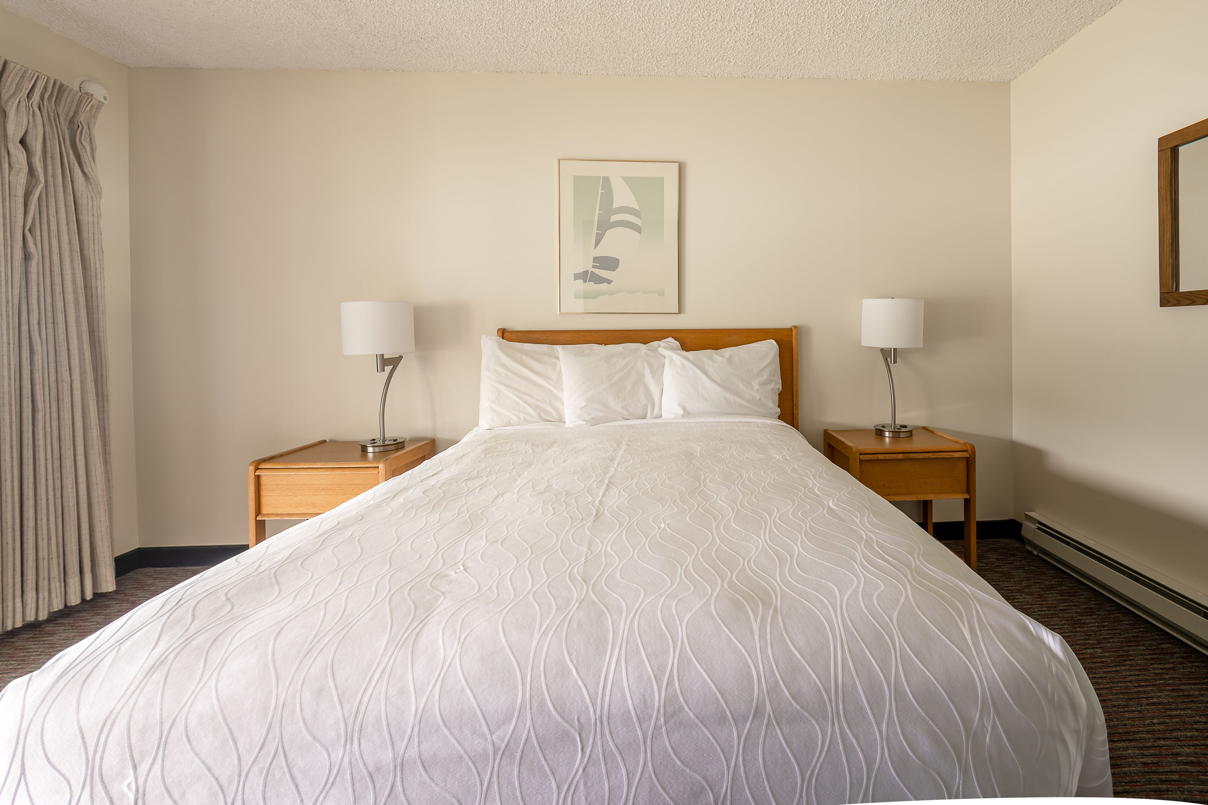A neatly made bed with a patterned bedspread, two bedside tables with lamps, a framed picture on the wall, and a window with curtains.