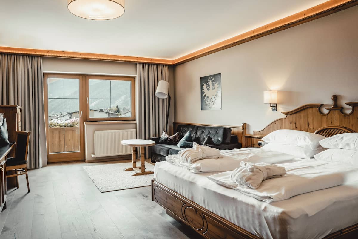 A hotel room with a bed, a desk, a chair, a window with curtains, a radiator, and a view of the mountains.
