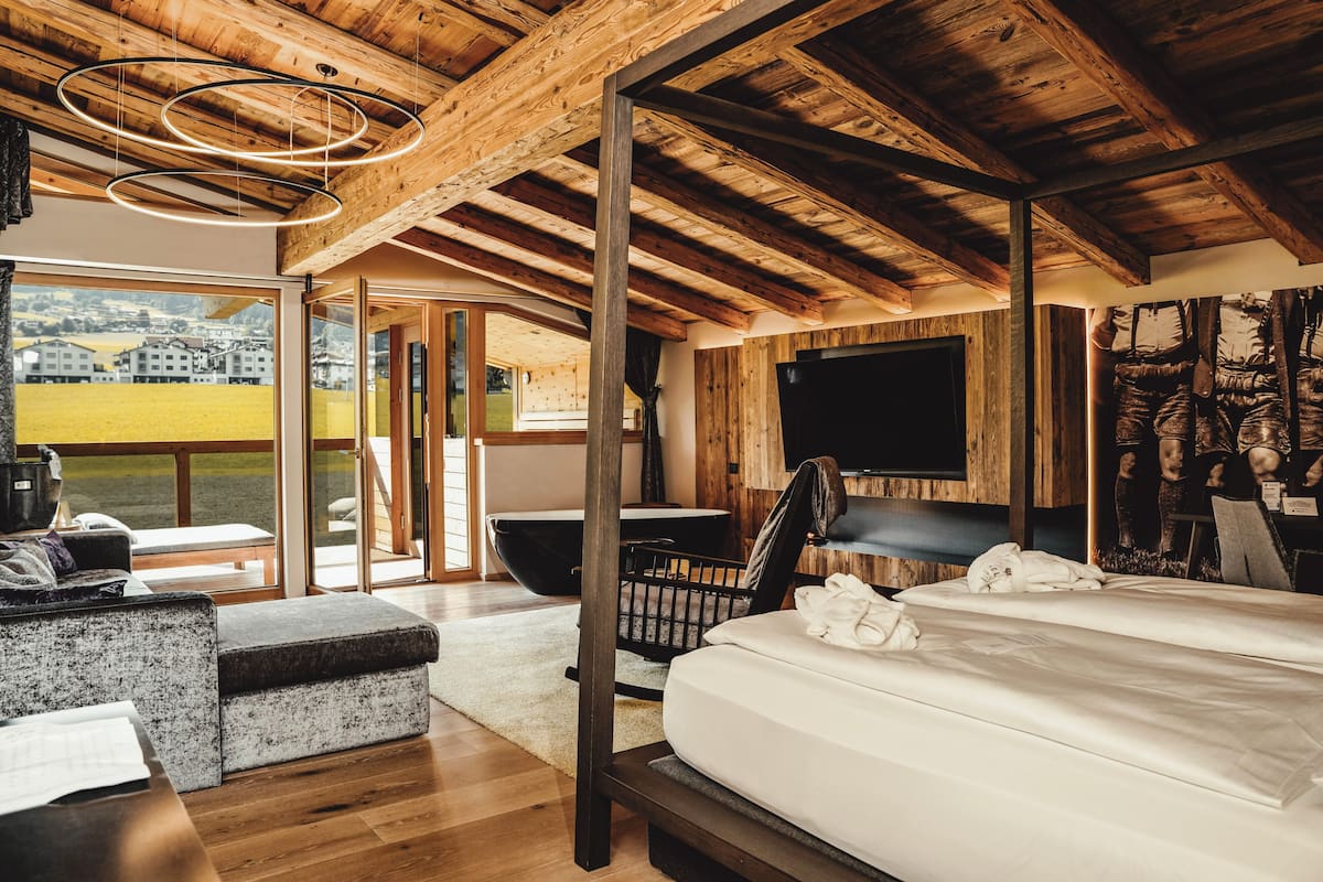 A modern bedroom with a large bed, wooden ceiling beams, a television, and a view of a green landscape.