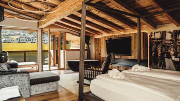 A modern bedroom with a large bed, wooden ceiling beams, a television, and a view of a green landscape.