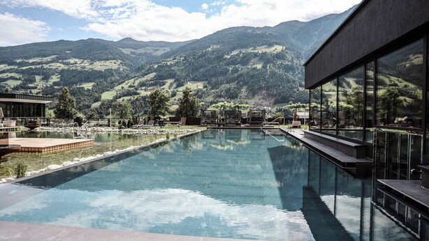 Una piscina con vistas a la montaña, un edificio moderno con grandes ventanales y una zona de terraza.