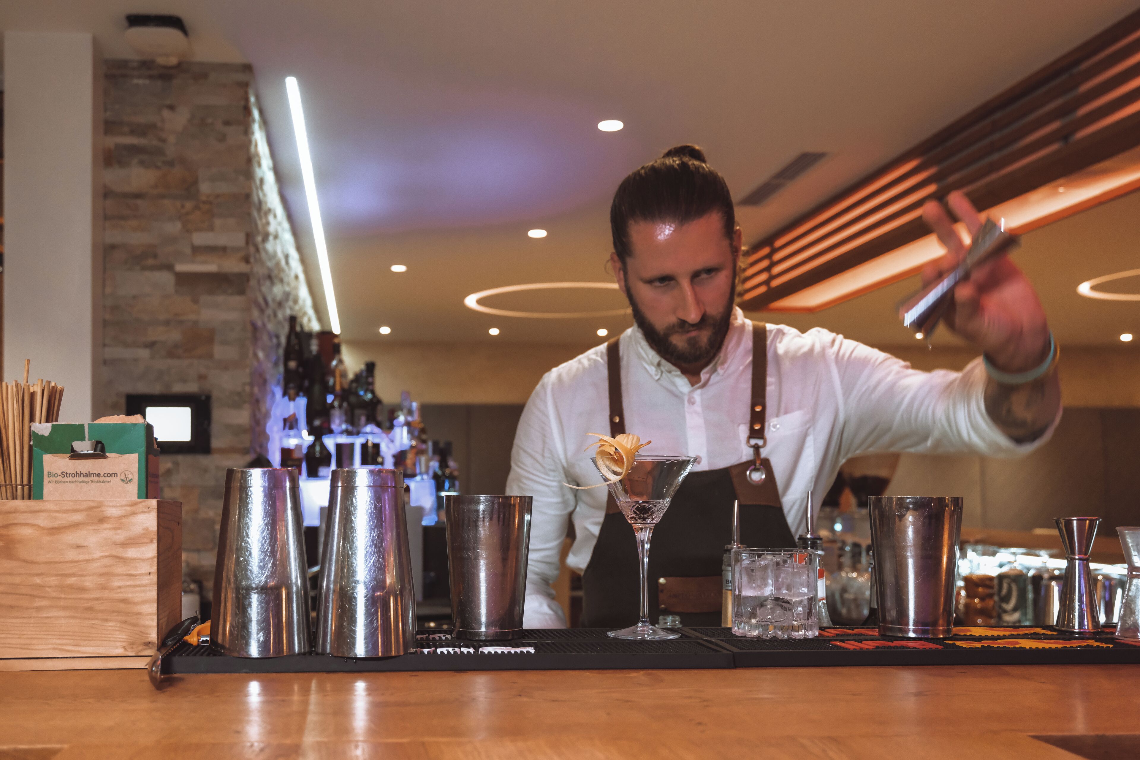 Un barista prepara un cocktail al bar, con un bancone in legno e shaker in metallo.
