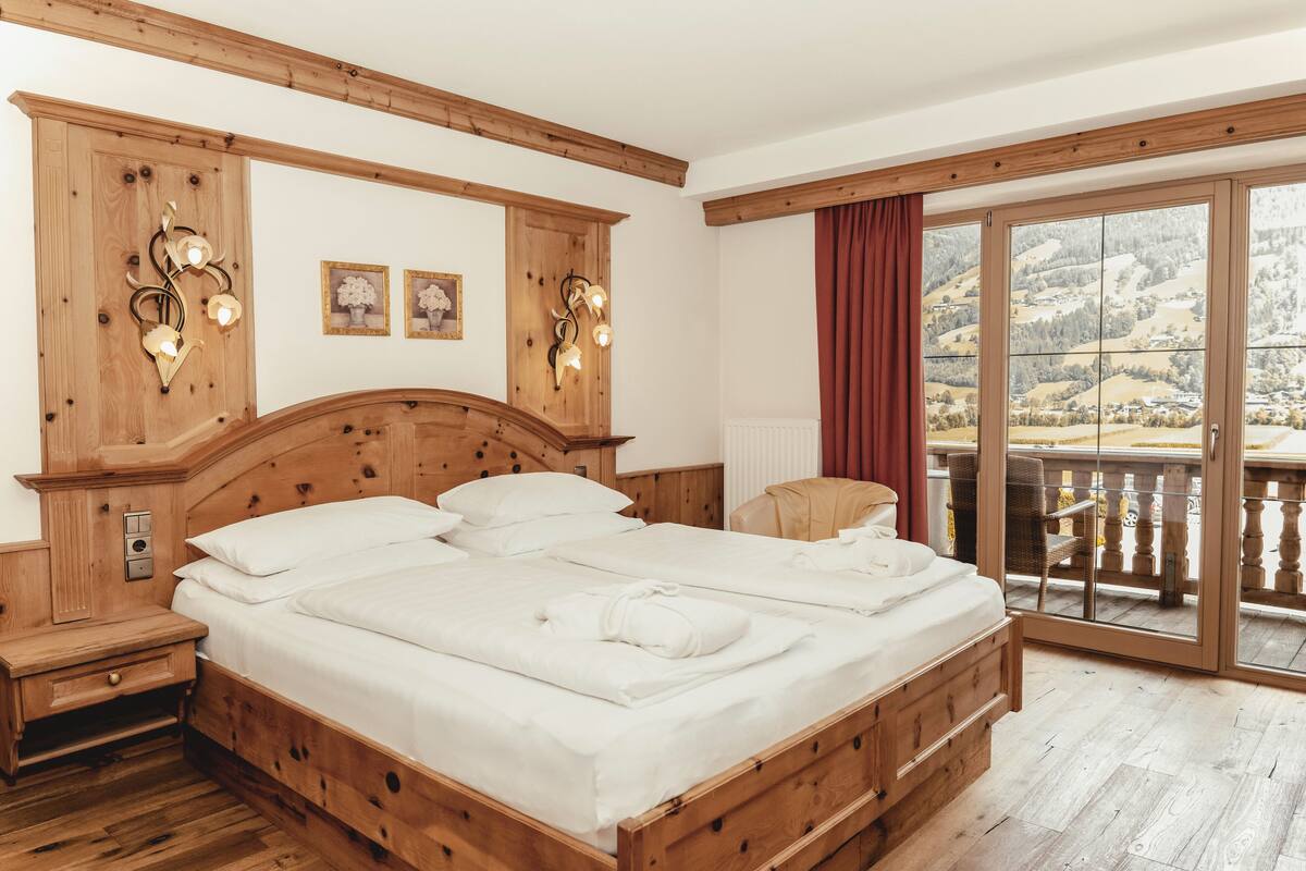 Un llit de fusta amb llençols blancs, una petita taula de nit de fusta, aplics de paret i vista a un balcó amb un paisatge idíl·lic.