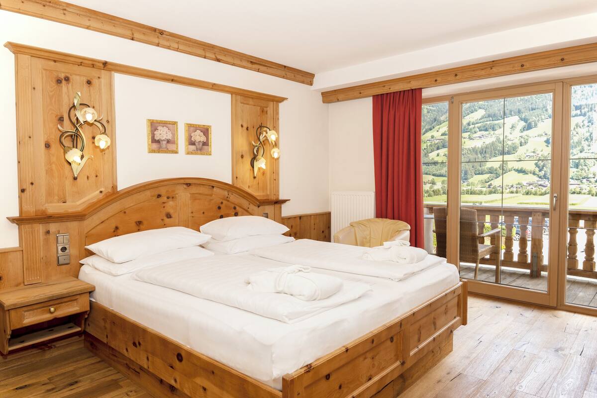 A wooden bed with white linens, a nightstand, wall sconces, and a view of a balcony with a red curtain.