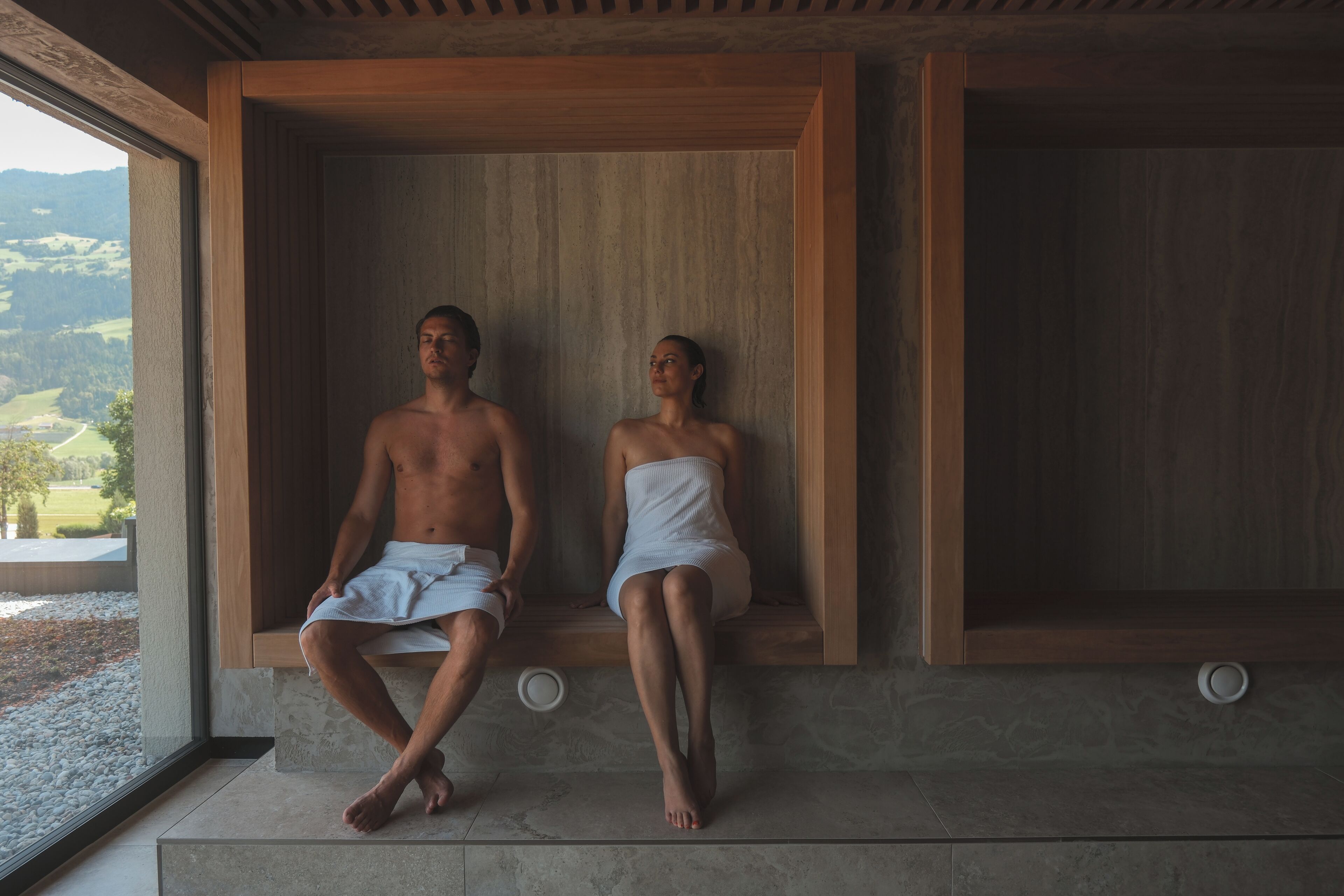 Two people sitting on a bench in a wooden cabin with a view of a landscape.