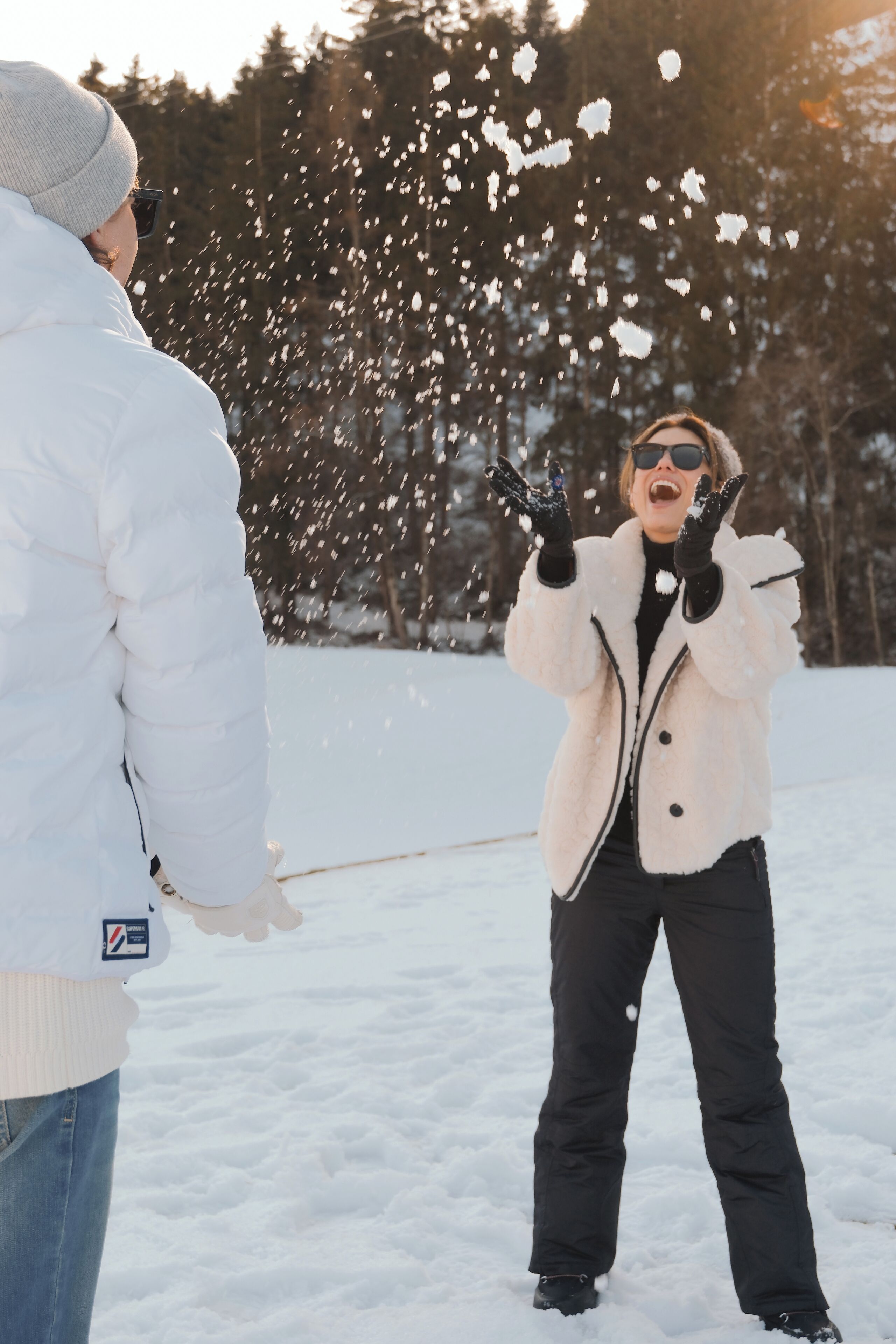 A person in a white jacket and black pants standing in a snowy area with trees in the background.