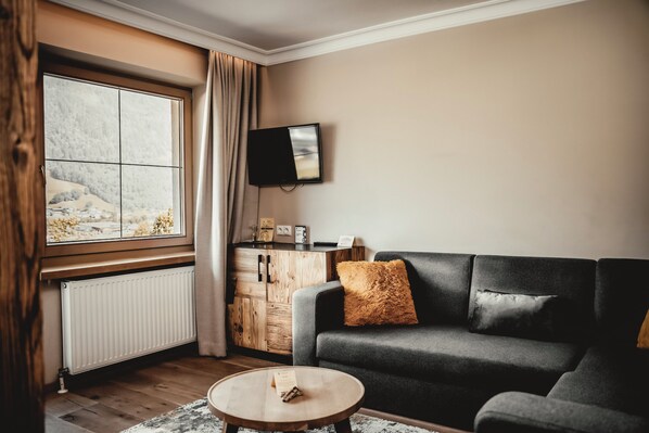 Una sala de estar moderna con un sofá gris, una mesa de centro de madera, un televisor montado en la pared y una ventana con vistas a un paisaje nevado.