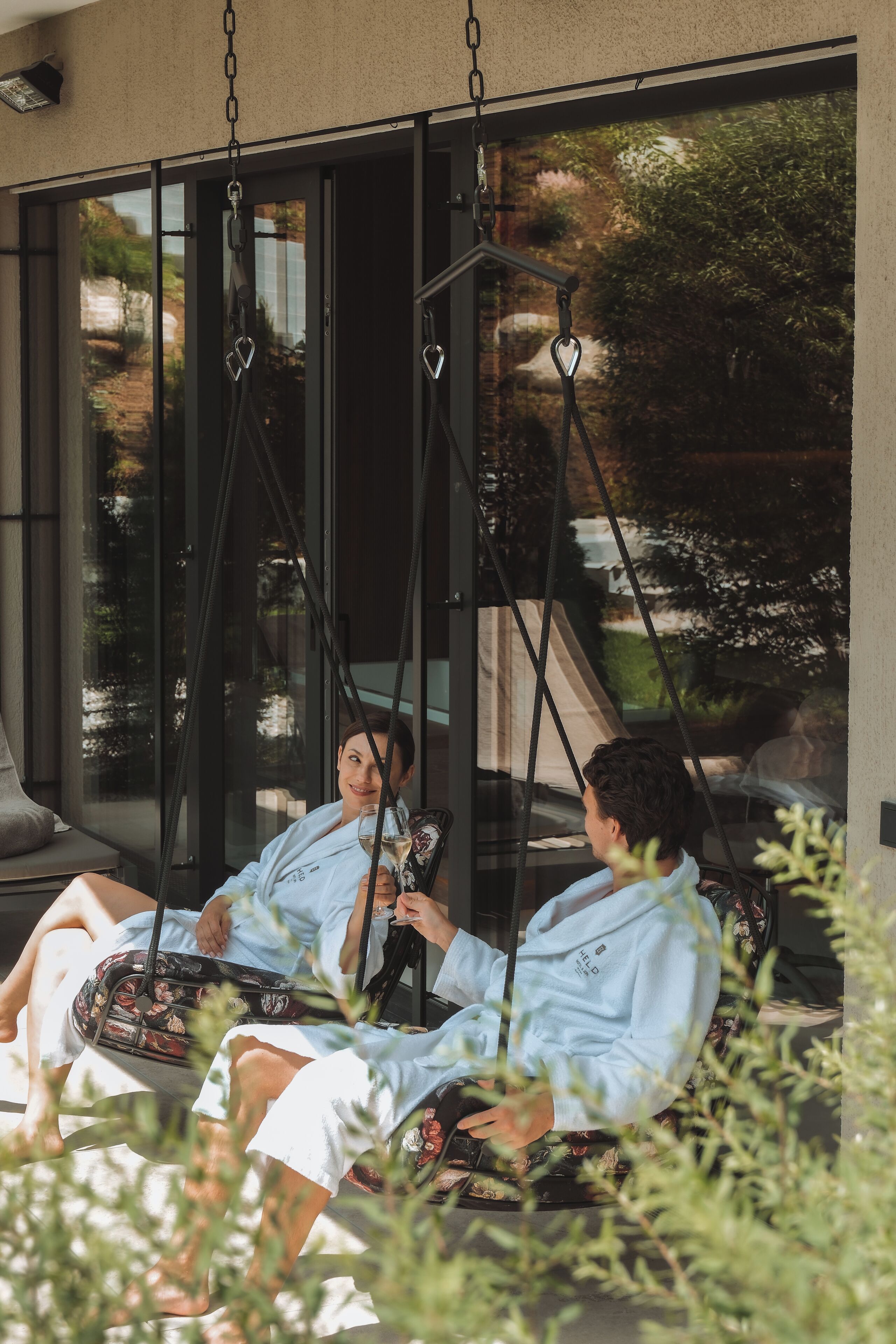 Dos personas sentadas en una mecedora al aire libre frente a un edificio con ventanas amplias.