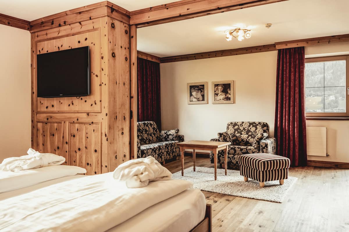 A hotel room with a wooden paneled wall, a flat-screen TV, two floral-patterned armchairs, a small wooden table, and a striped ottoman.