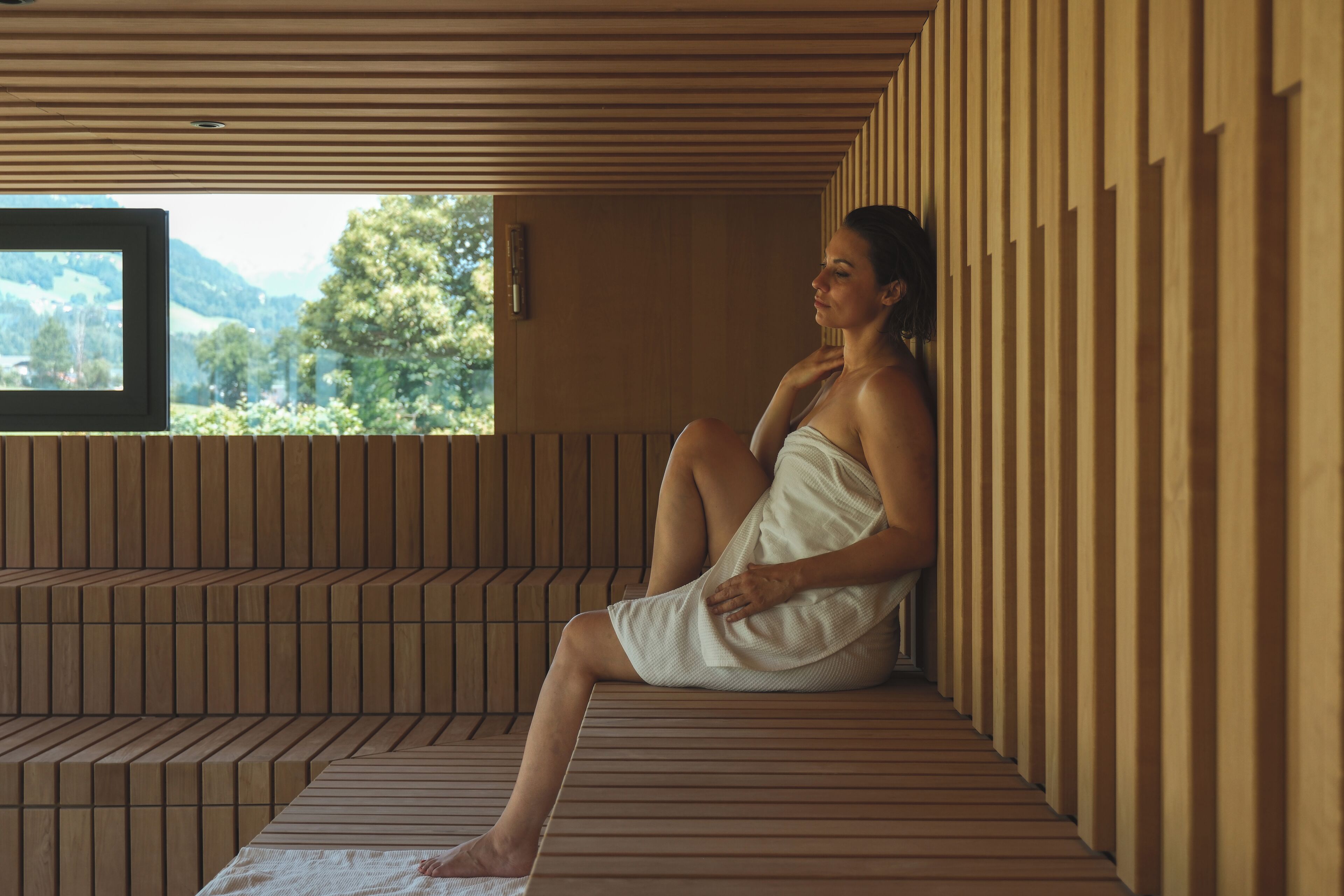 A person sitting on a wooden bench in a room with a view of greenery and hills.