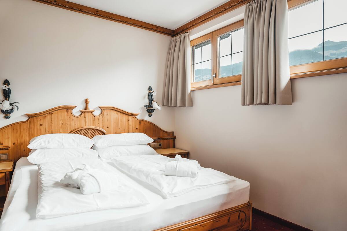 A neatly made bed with white linens and a wooden headboard, a bedside table with a lamp, and a window with curtains.