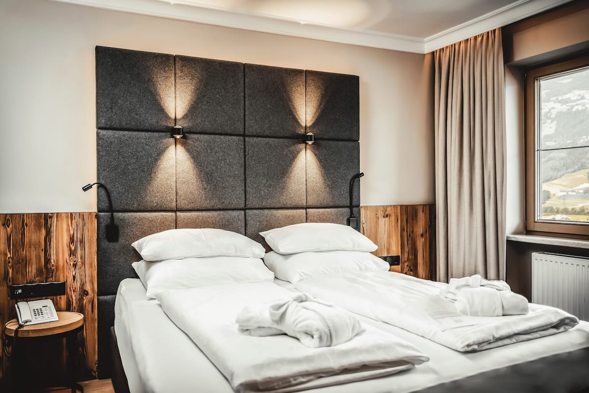 A neatly made bed with white linens, a headboard with illuminated panels, a bedside table with a lamp, and a window with curtains.