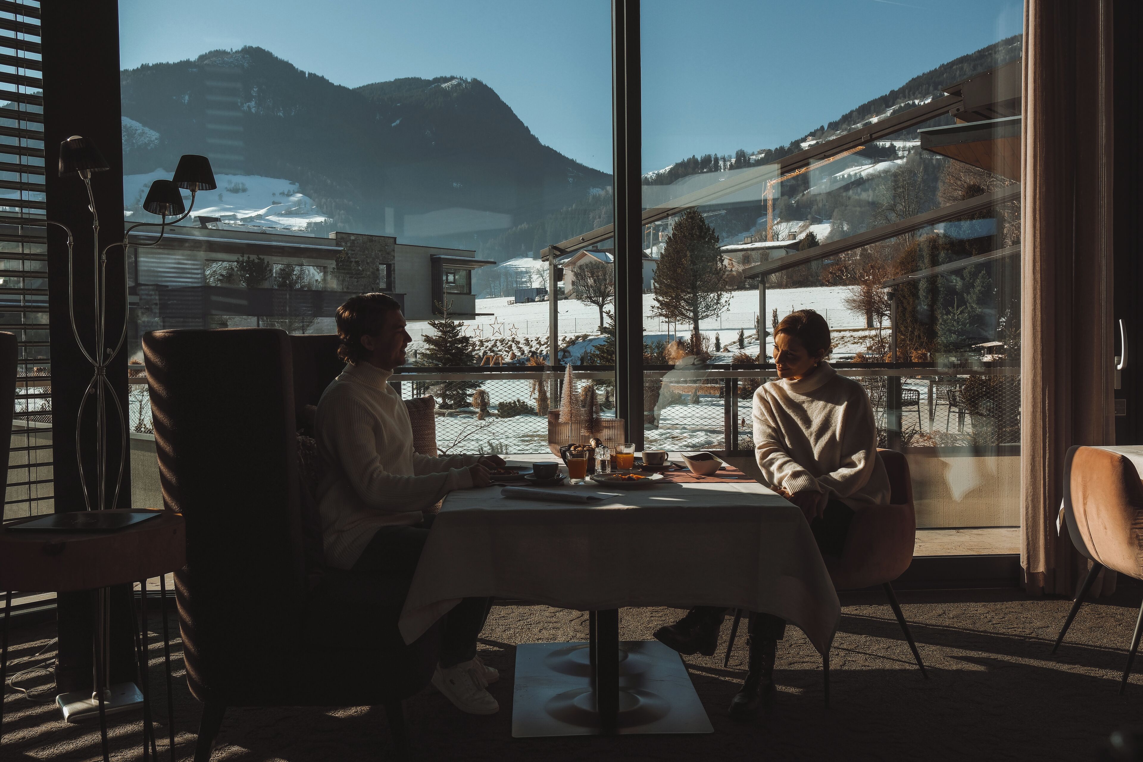 Dos personas sentadas en una mesa con vista a un paisaje montañoso nevado a través de amplios ventanales.