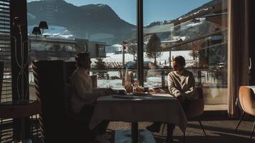 Dos personas sentadas en una mesa con vista a un paisaje montañoso nevado a través de amplios ventanales.