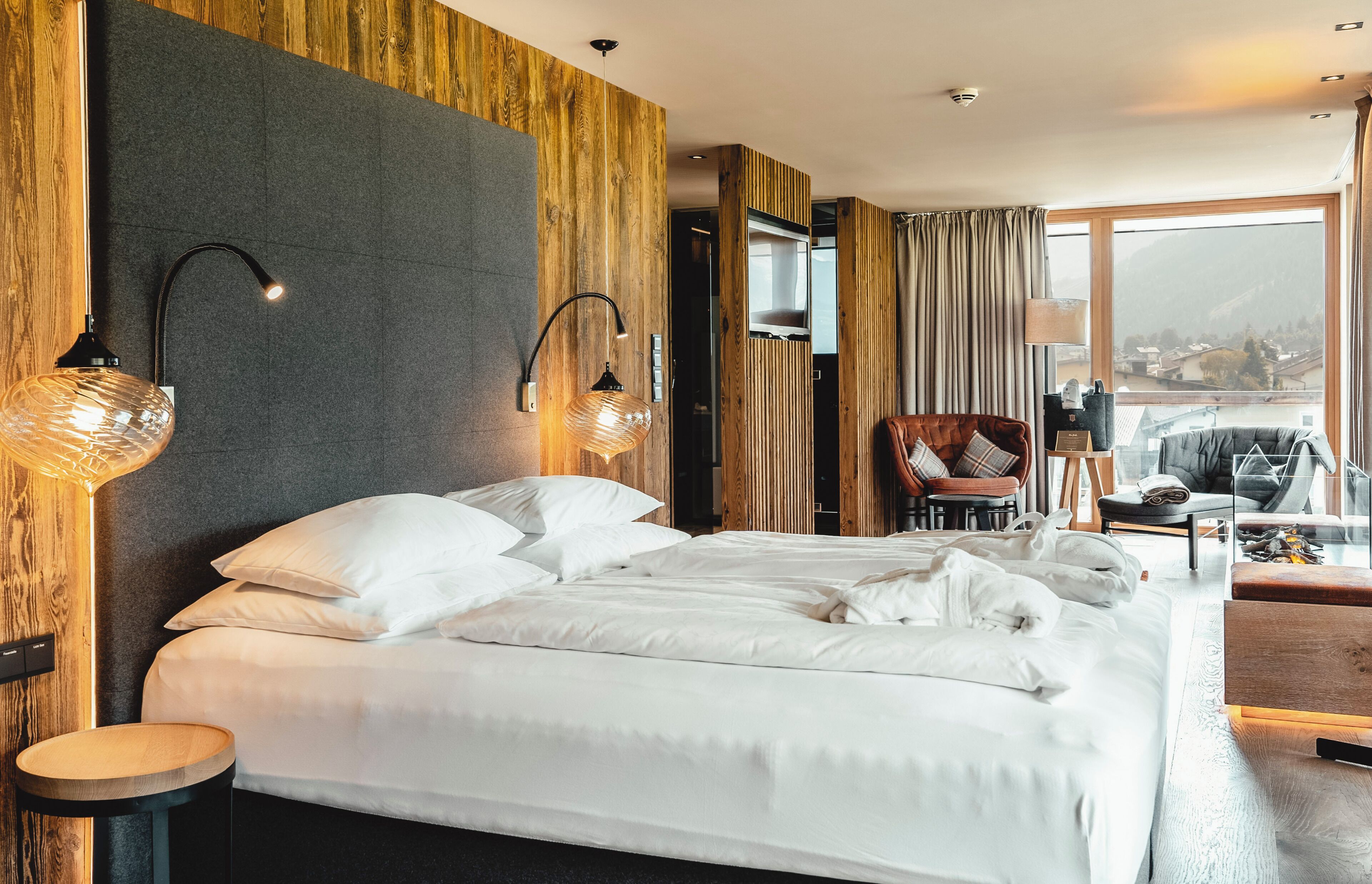 A modern hotel room with a large bed, two bedside lamps, a TV, a seating area with a chair and table, and a view of the outdoors.
