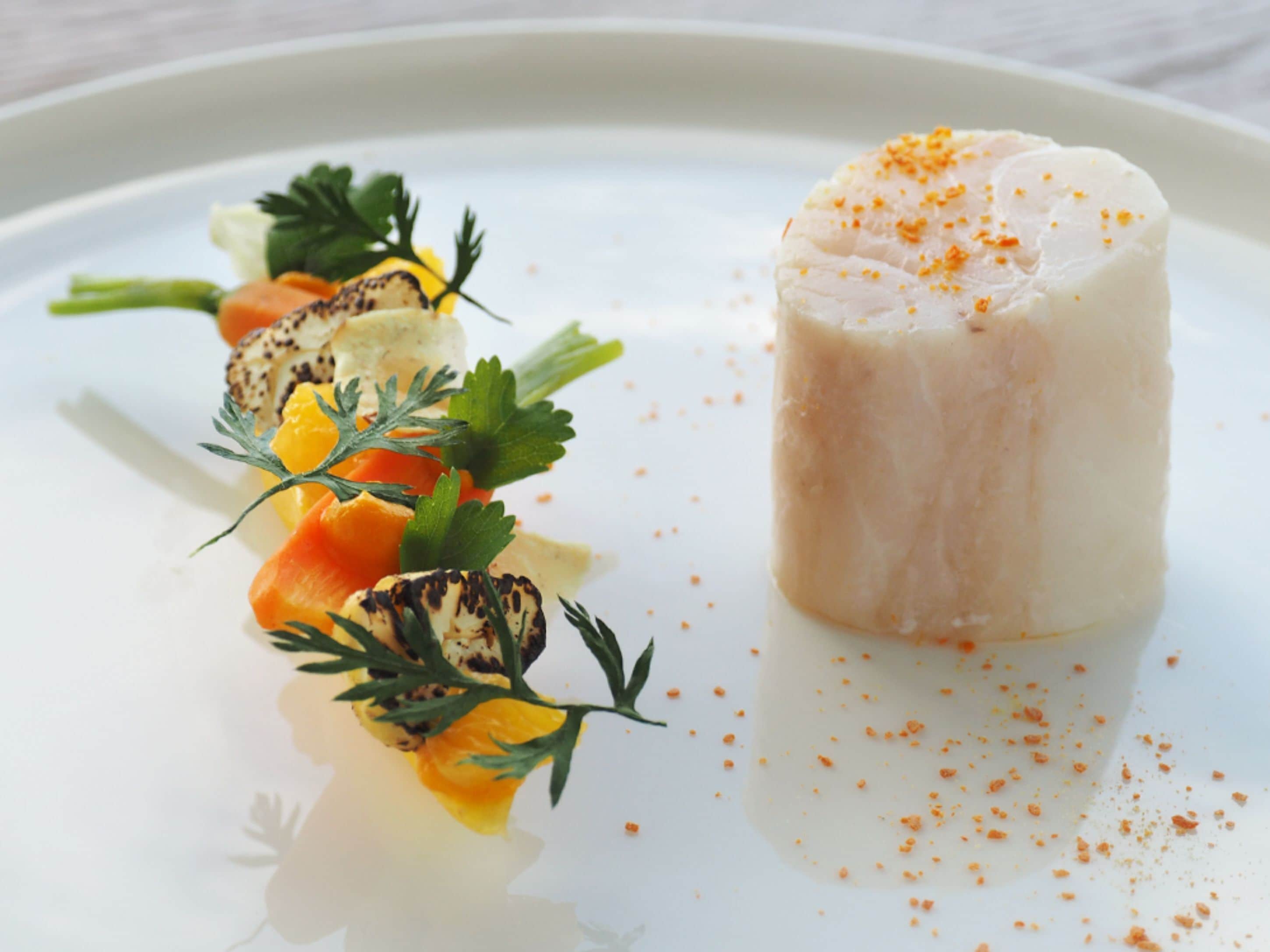 A white plate with a cylindrical food item, possibly a type of cheese or terrine, garnished with orange powder and surrounded by vegetables like carrots and herbs.