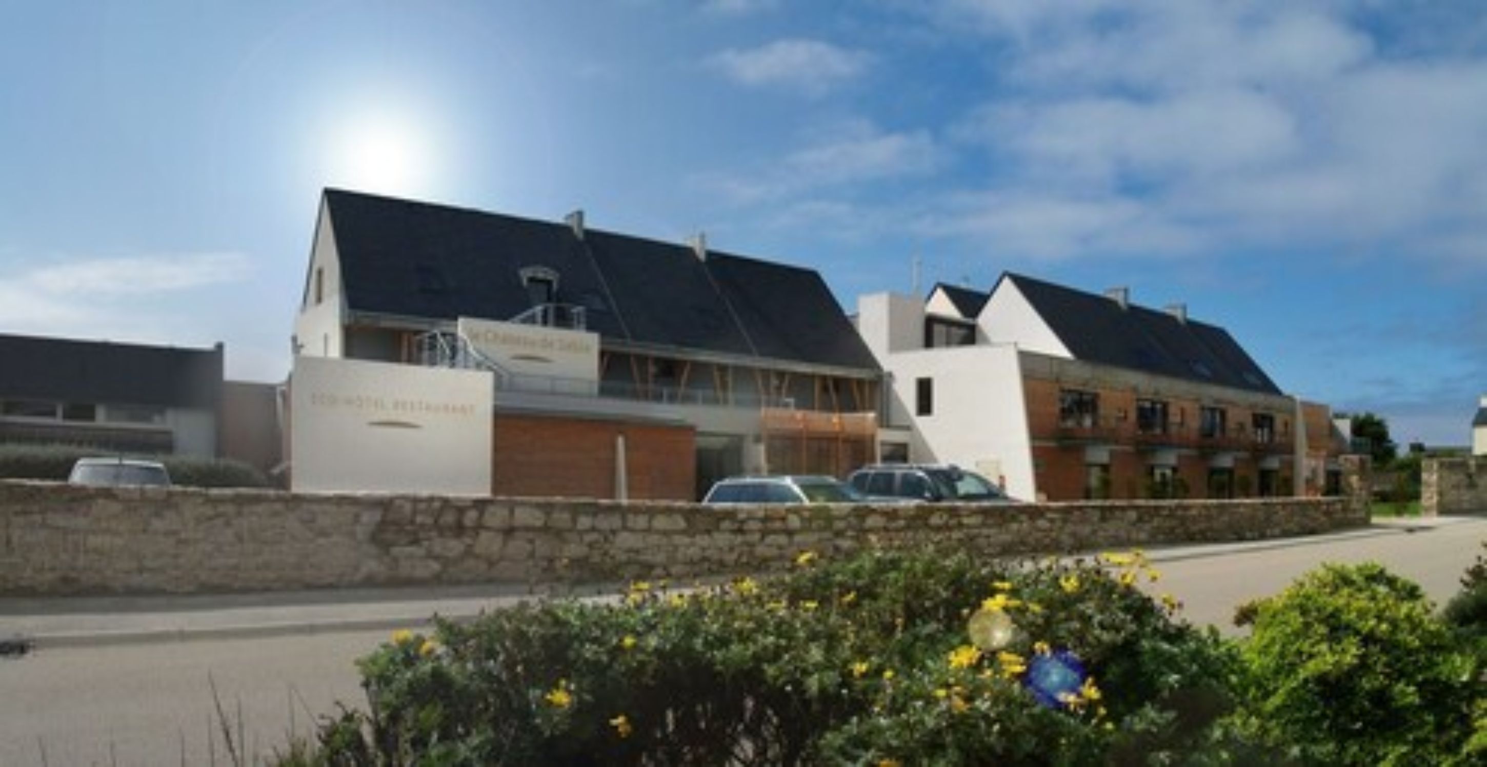 A modern building with a sign that reads "Hotel de la Mer." It features a stone wall in front and a parking area with cars.