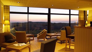 Un moderno vestíbulo de hotel con una vista panorámica del paisaje urbano al atardecer, amueblado con sofás de cuero, mesas de madera y una recepción.