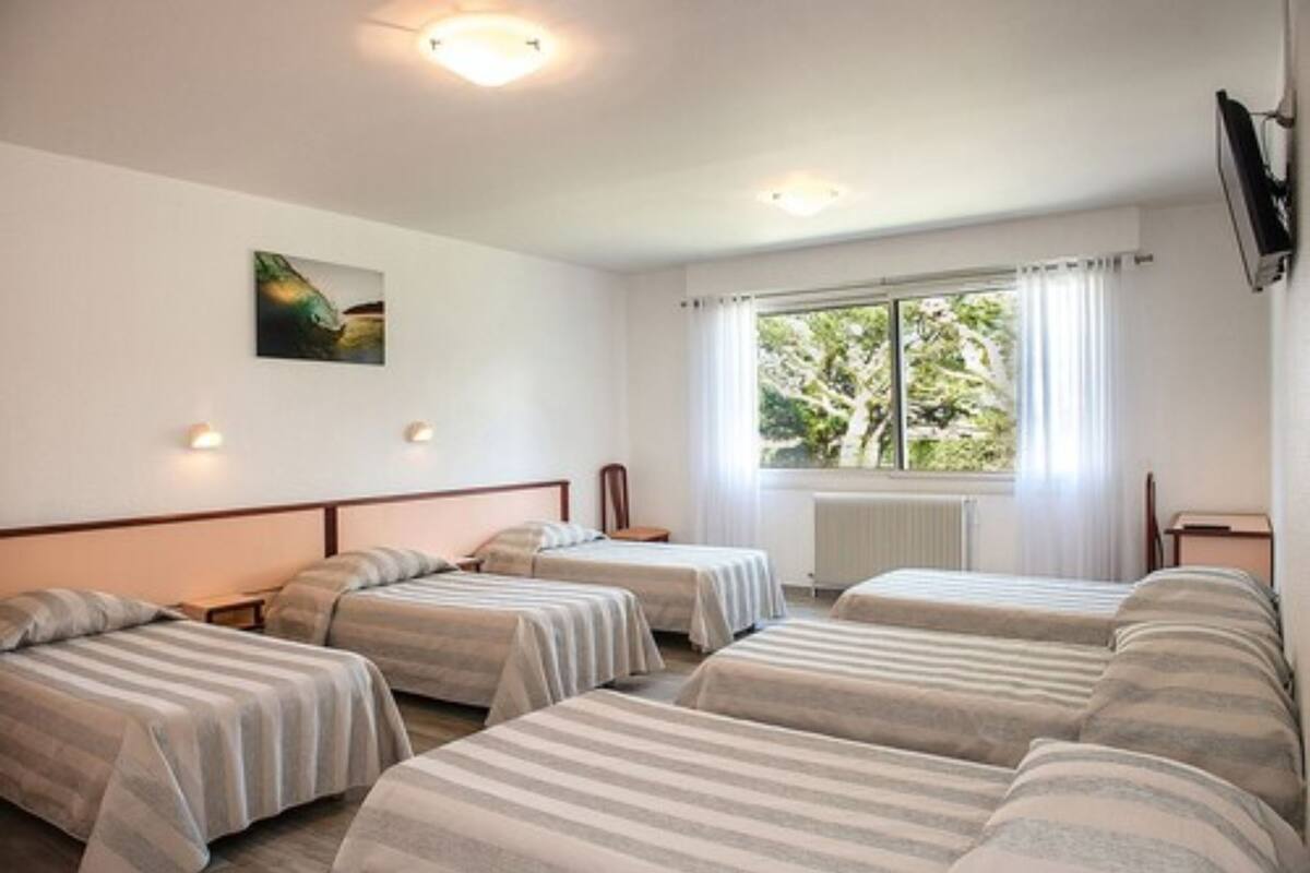 Camera d'albergo con quattro letti singoli, un'ampia finestra con tende, un quadro appeso al muro e un televisore a parete.