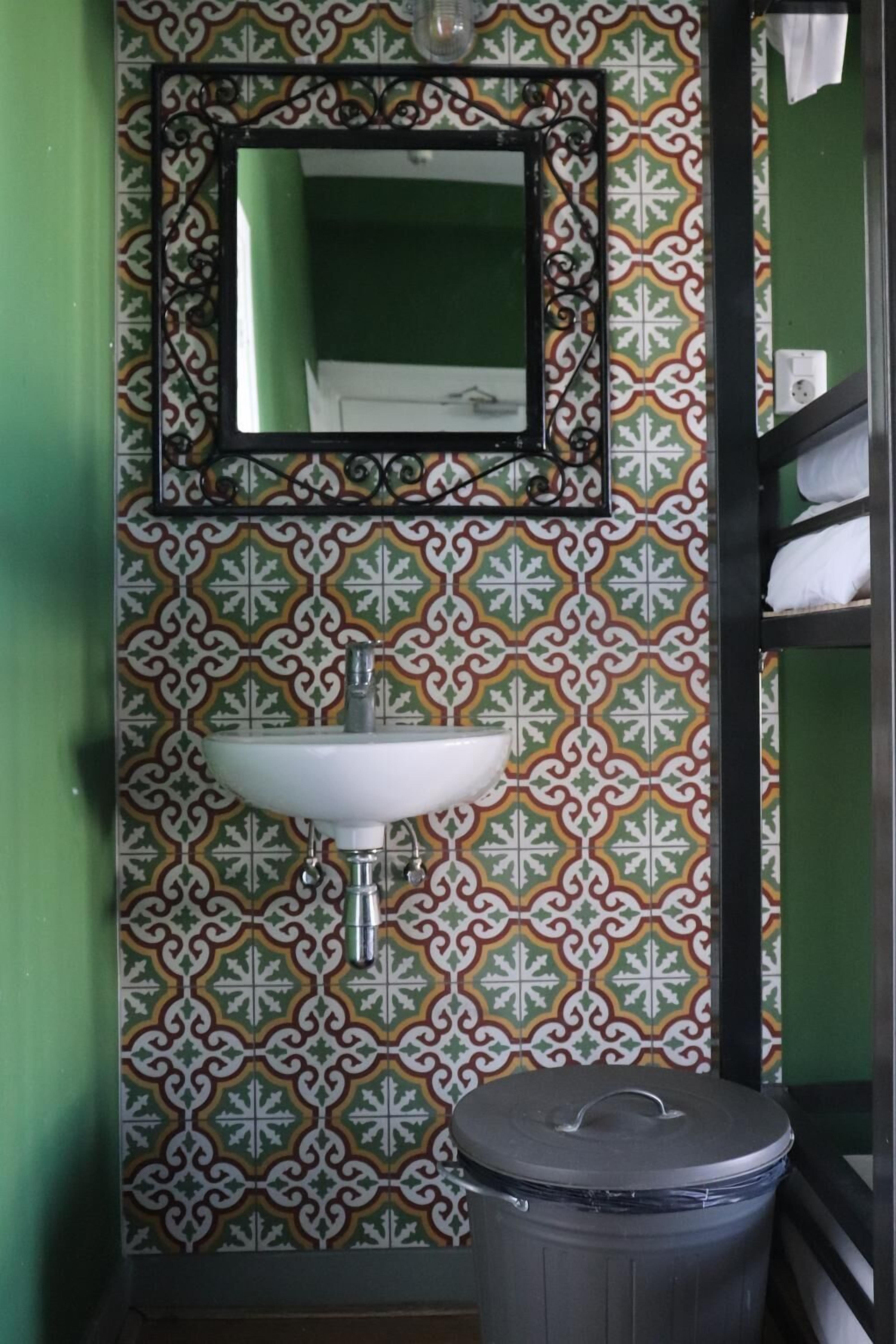 A bathroom with a patterned tile wall, a white sink, a mirror, and a trash bin.