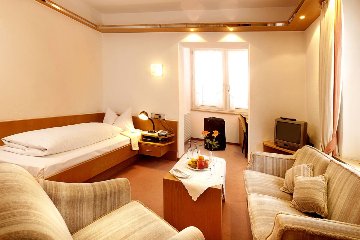 A hotel room with a bed, a sofa, a small table with a vase and fruit, a TV, and a window with curtains.