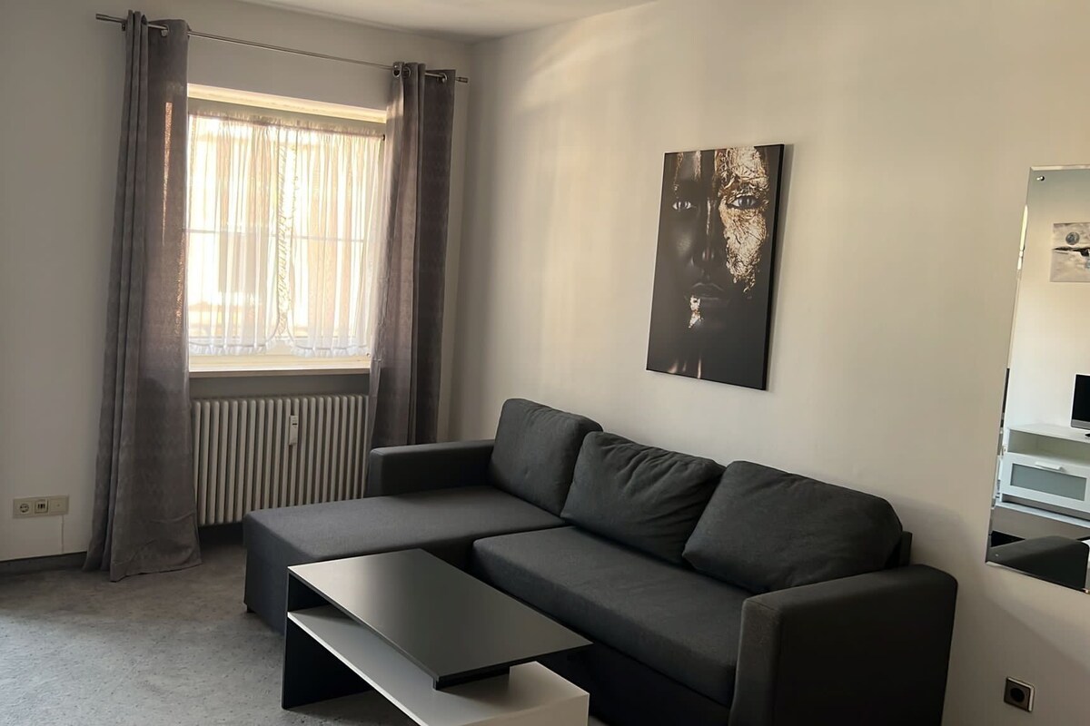 A modern living room with a grey sofa, a black coffee table, and a large window with sheer curtains.