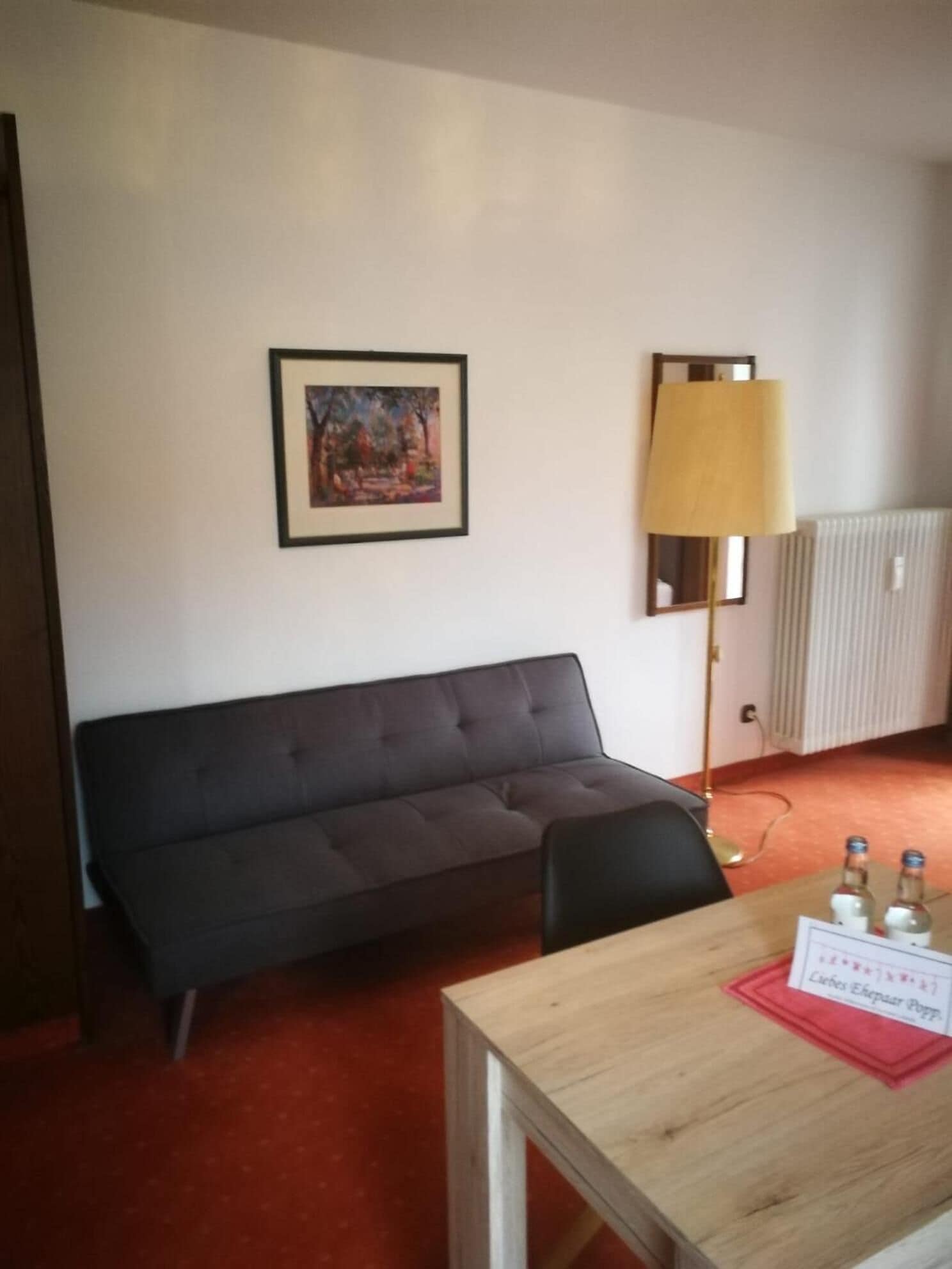 A room with a dark grey sofa, a wooden table with a red placemat, a framed painting on the wall, and a floor lamp.