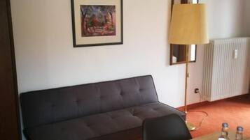 A room with a dark grey sofa, a wooden table with a red placemat, a framed painting on the wall, and a floor lamp.