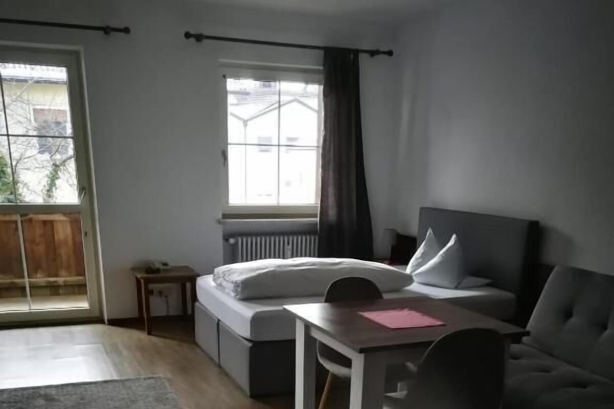 A modern bedroom with a bed, a desk, and a chair. There is a window with curtains and a view of buildings outside.