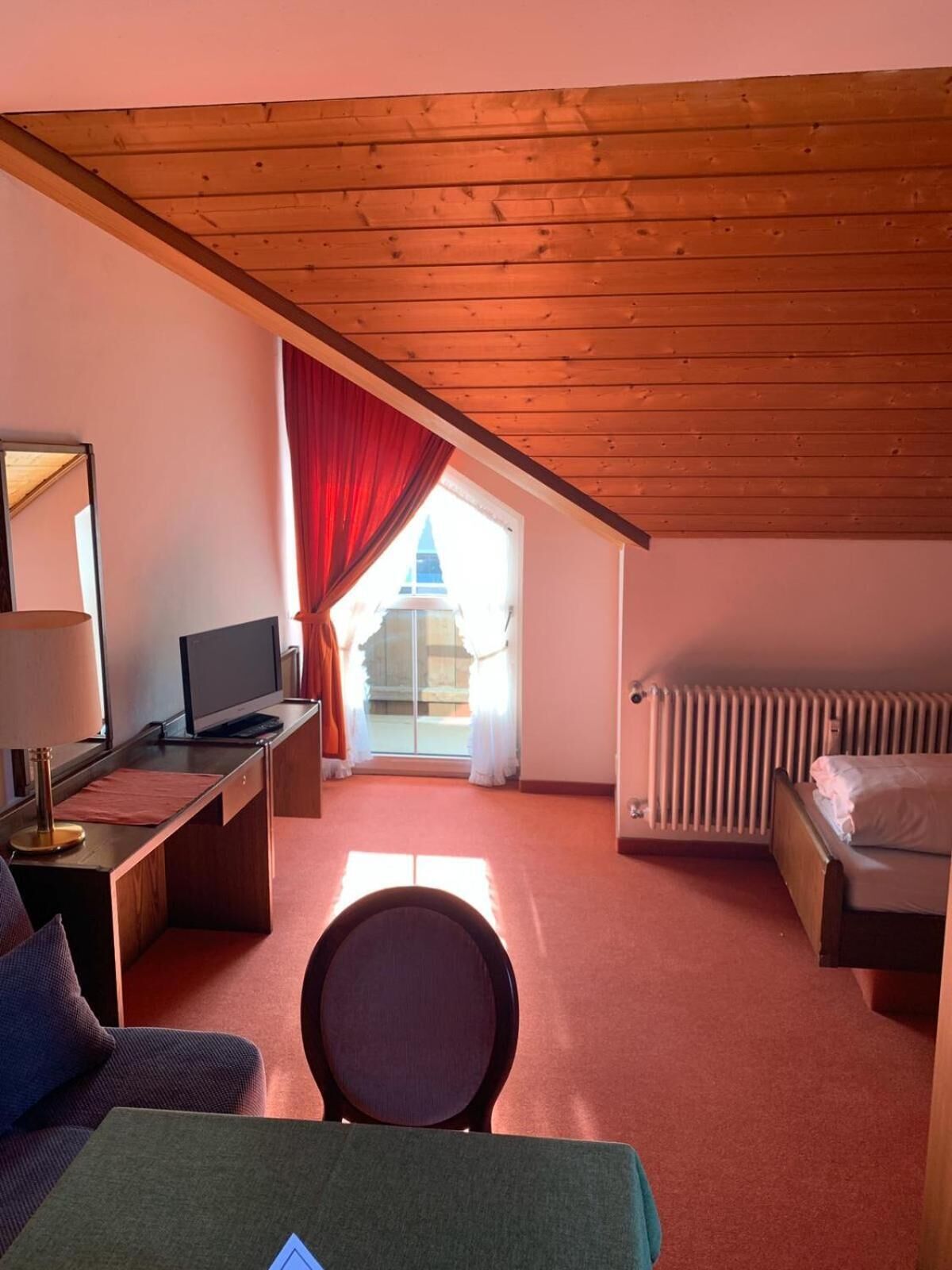 A small, cozy room with a wooden ceiling, a desk with a computer, a bed, and a radiator.