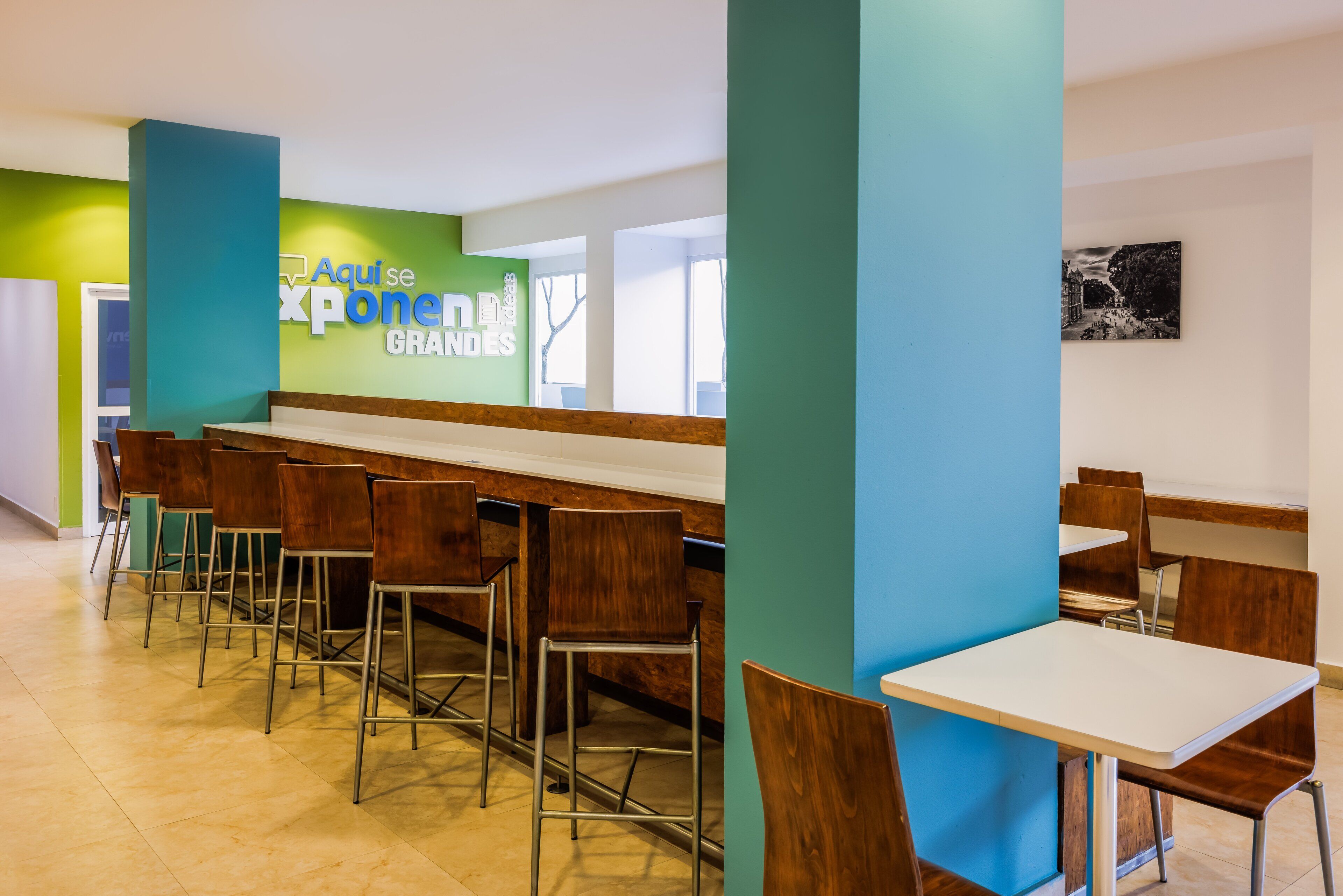 A cafeteria with high tables and chairs, a green accent wall, and a sign reading "Aqui se Expone Ideas".