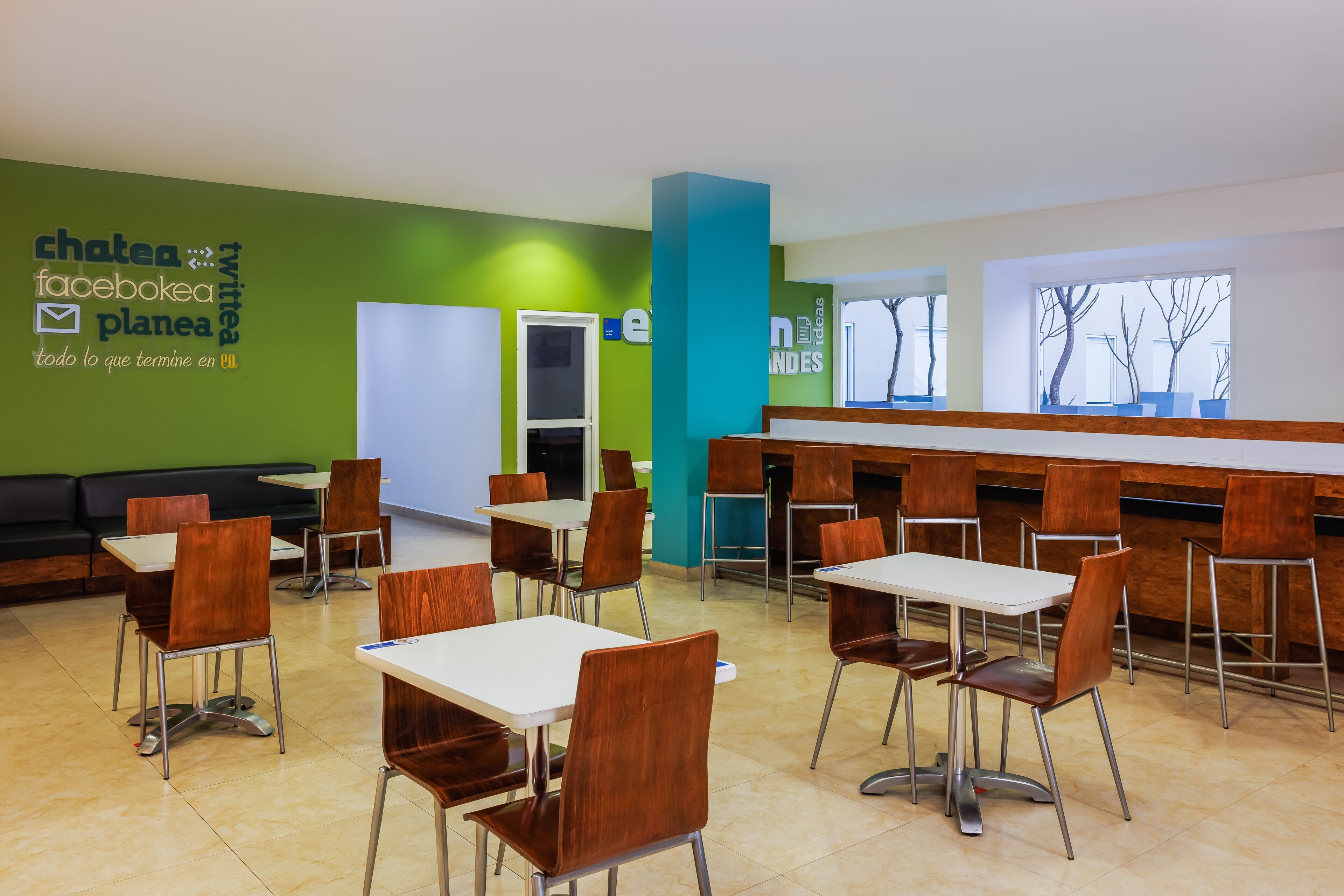 A modern dining area with tables and chairs, a green wall with logos, and a window with bare trees.