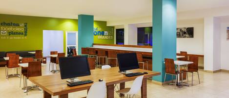 A modern library with wooden tables and white chairs, featuring a green wall with social media logos and a blue pillar.