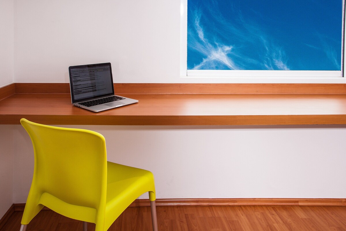 A laptop on a wooden shelf, a yellow chair, and a window with a blue sky view.