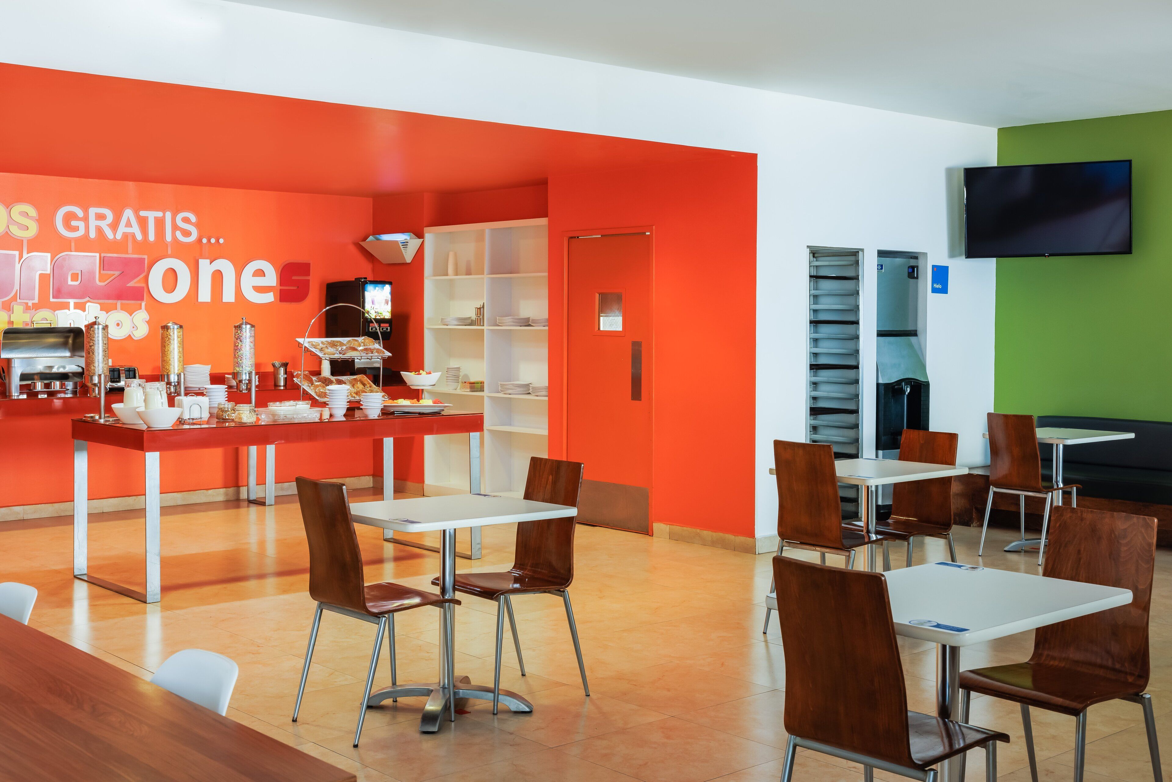 Un comedor moderno con una pared de color naranja brillante, mesas blancas y sillas marrones.