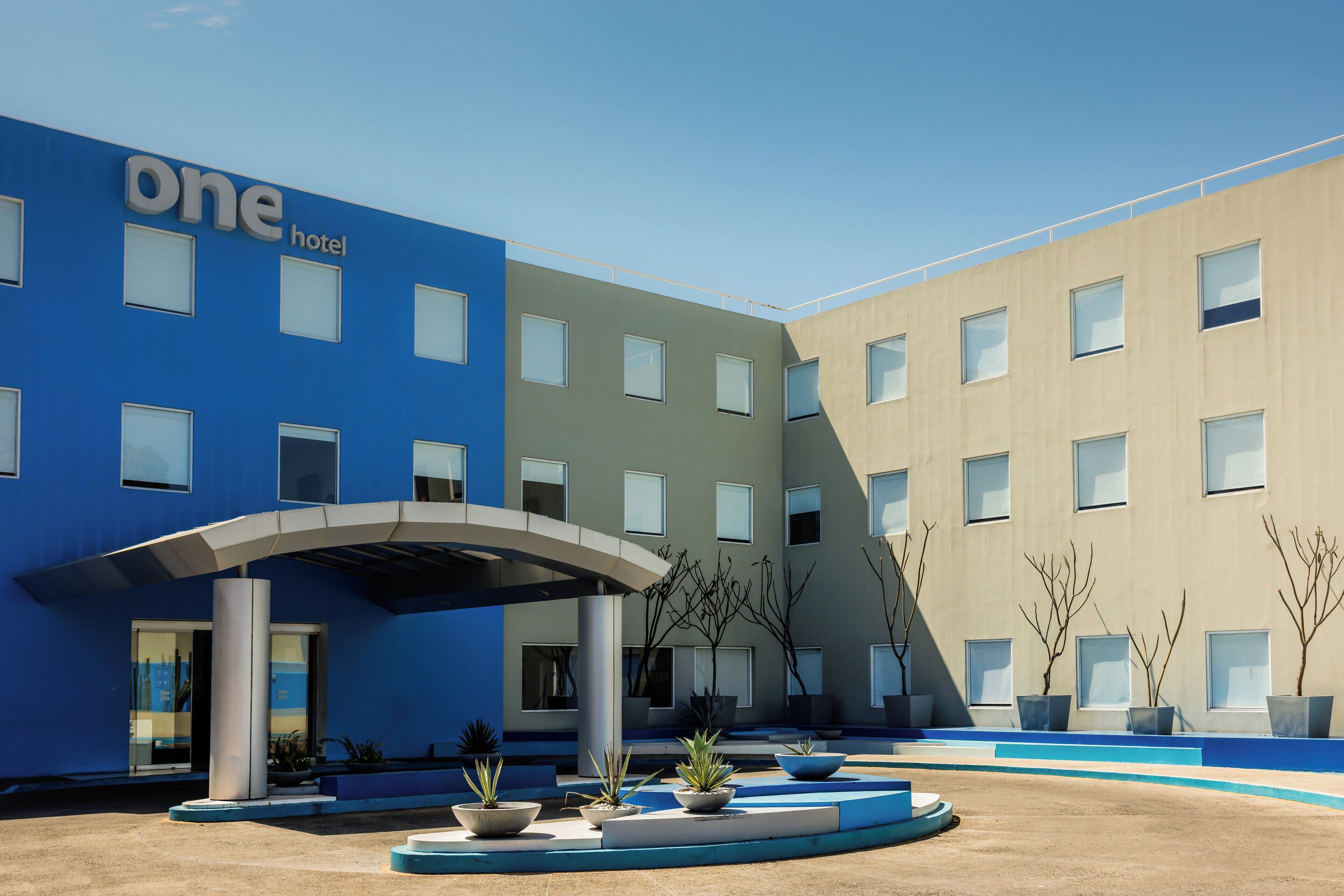 Un hotel moderno con fachada azul y color beige, un acceso cubierto y un patio ajardinado con macetas.