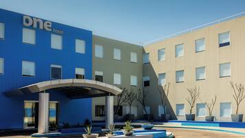 Un hotel moderno con fachada azul y color beige, un acceso cubierto y un patio ajardinado con macetas.
