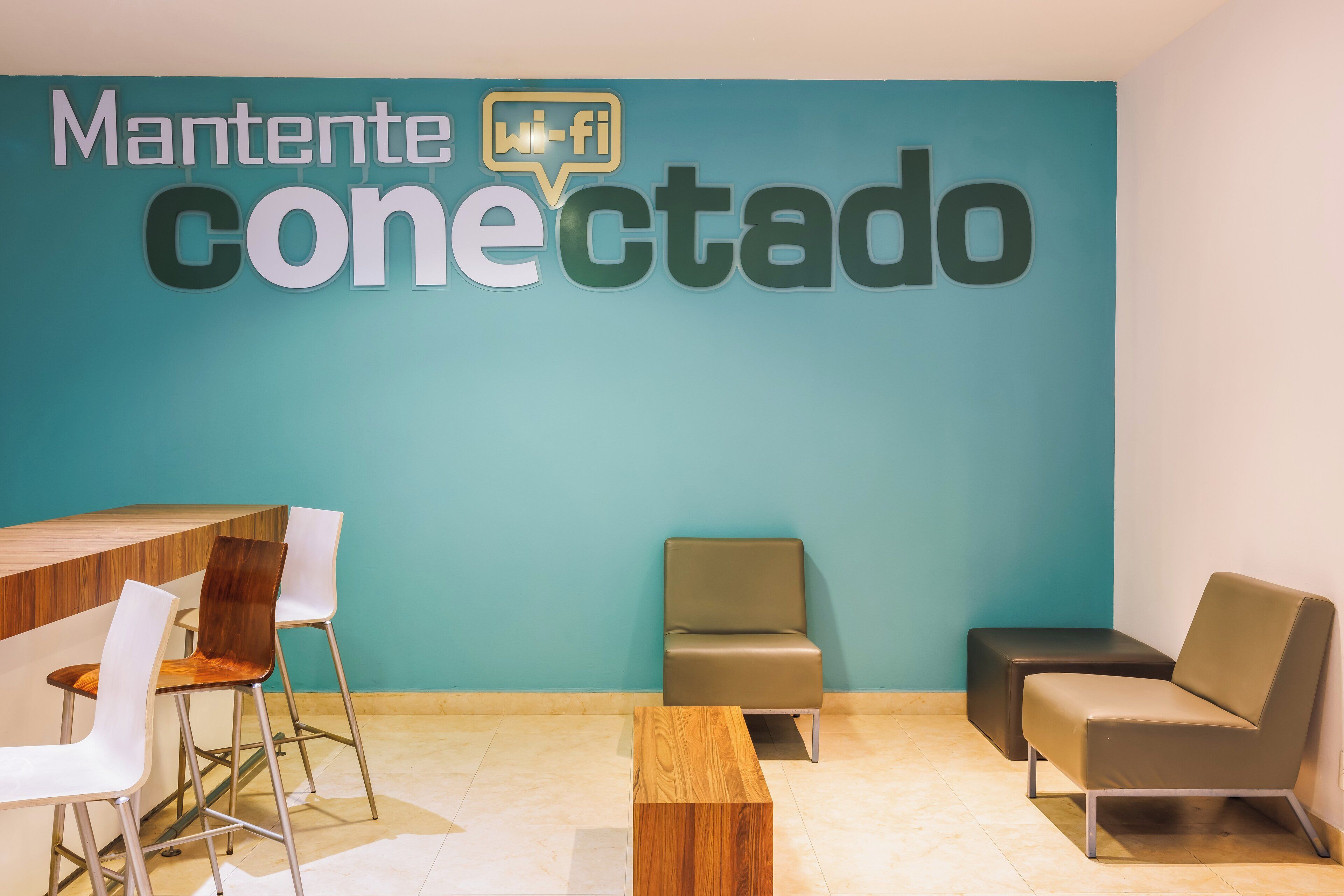 Un área de espera moderna con una pared azul verdoso, una mesa de madera, dos sillas blancas, un sofá marrón y un pequeño otoman marrón.