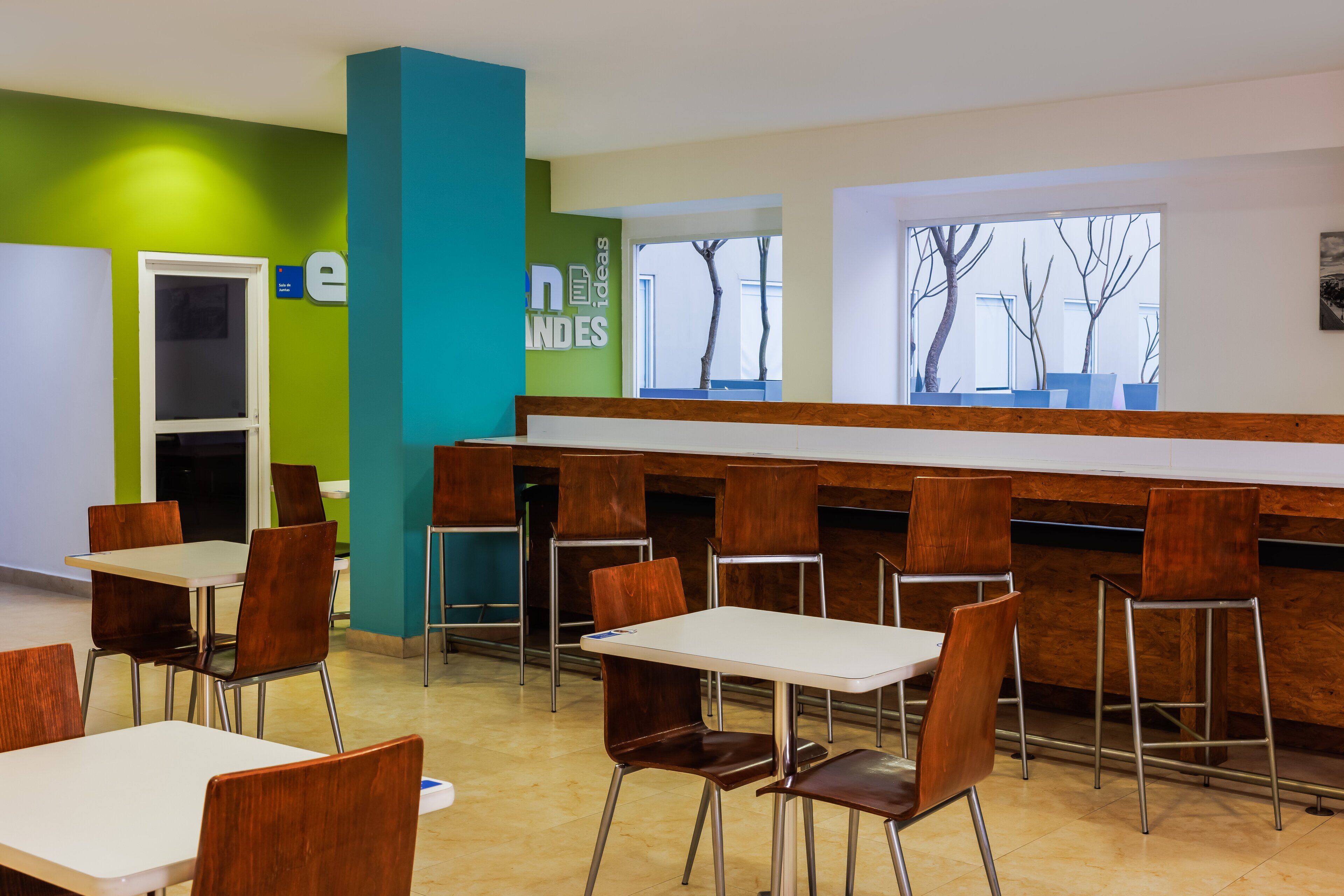 A modern cafeteria with white tables and brown chairs, green and blue walls, and large windows with a view of bare trees.