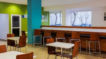 A modern cafeteria with white tables and brown chairs, green and blue walls, and large windows with a view of bare trees.
