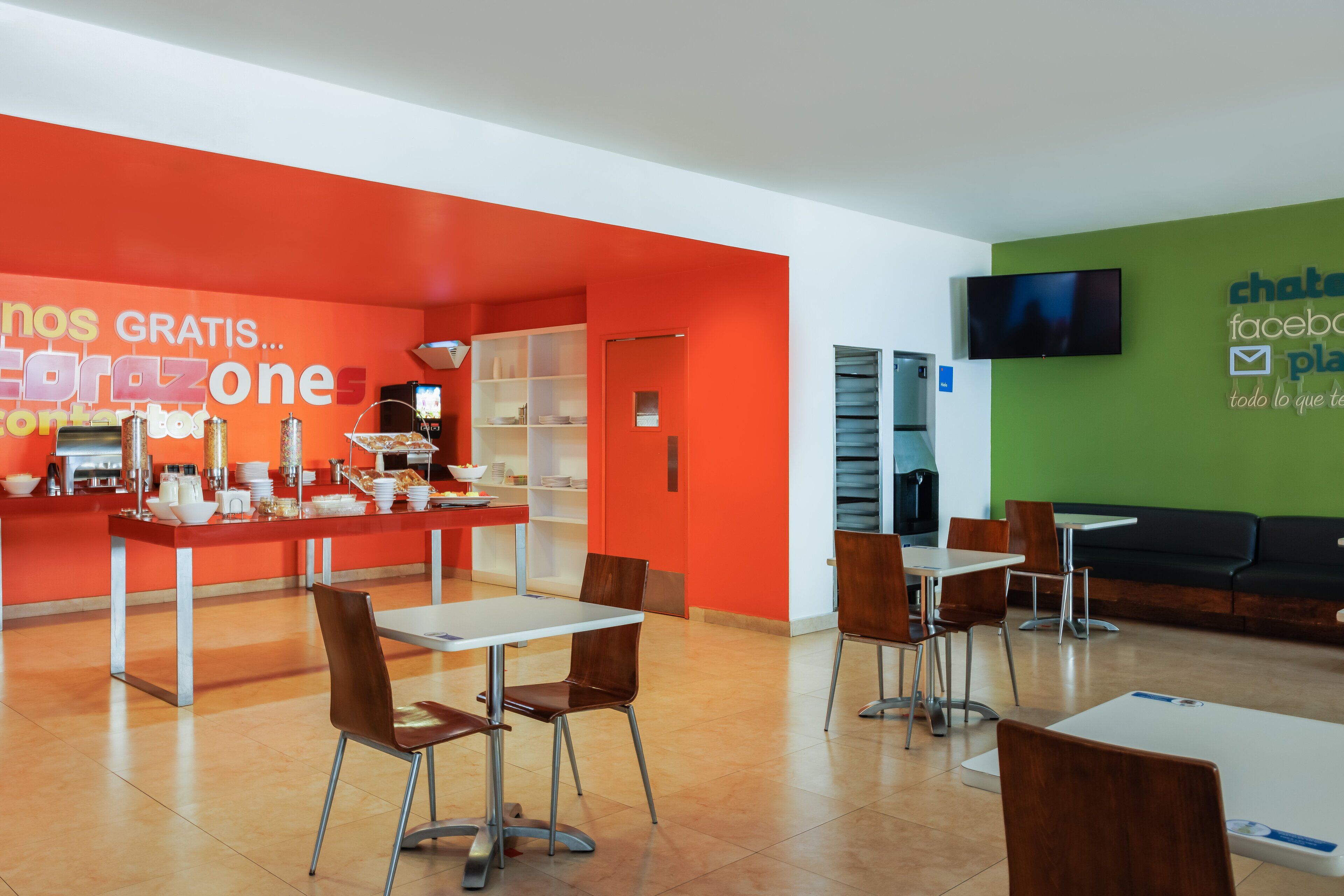 Una cafetería moderna con una pared de color naranja brillante, mesas blancas y sillas marrones.