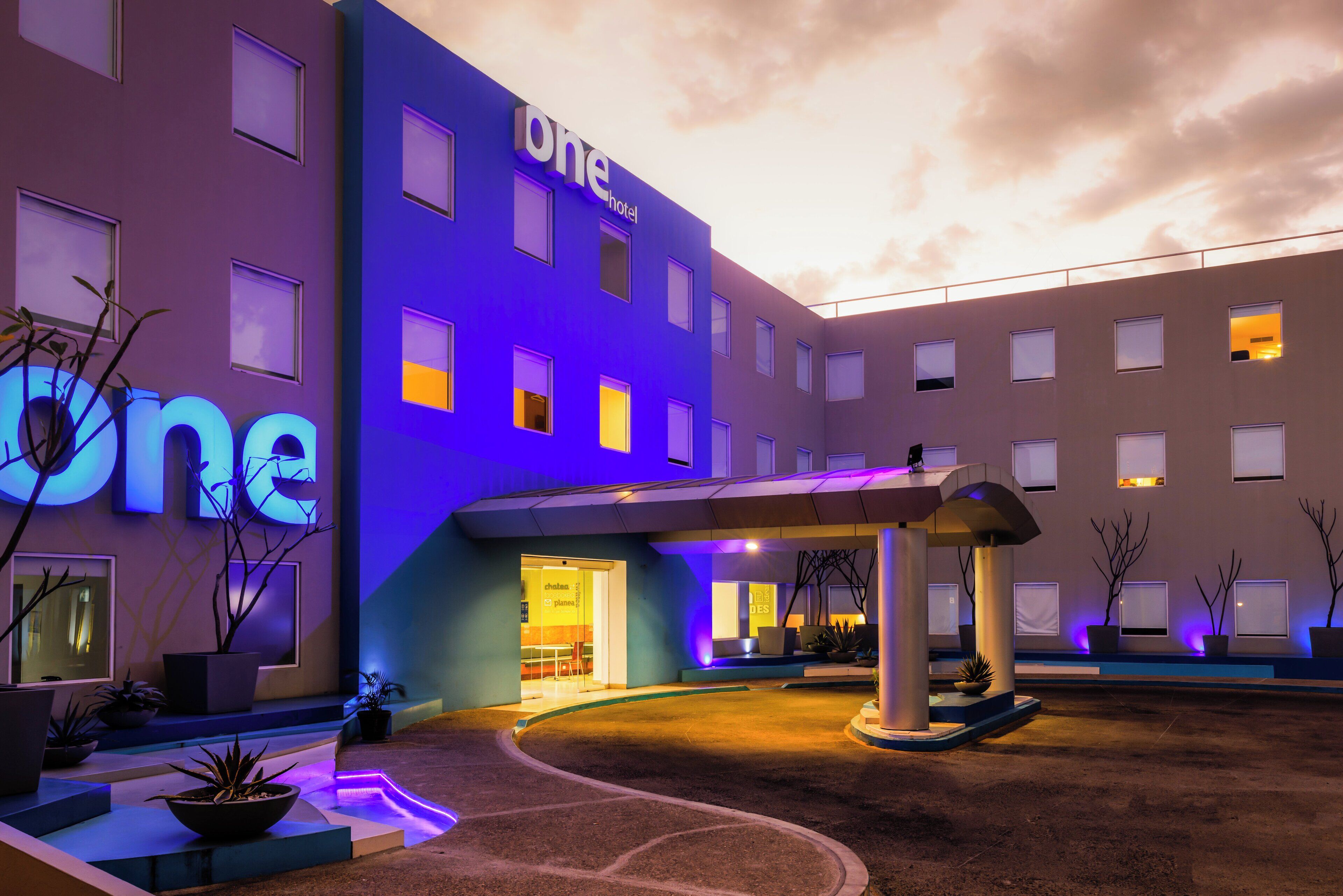 Un hotel de diseño moderno, con un llamativo letrero que dice 'one' en la fachada, acceso cubierto y un área exterior ajardinada con plantas en macetas.