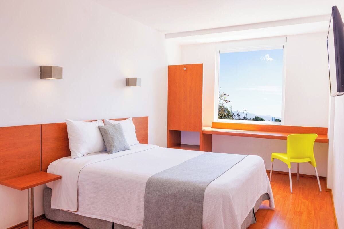 A hotel room with a bed, a desk, a chair, a window with a view, and a wall-mounted light.