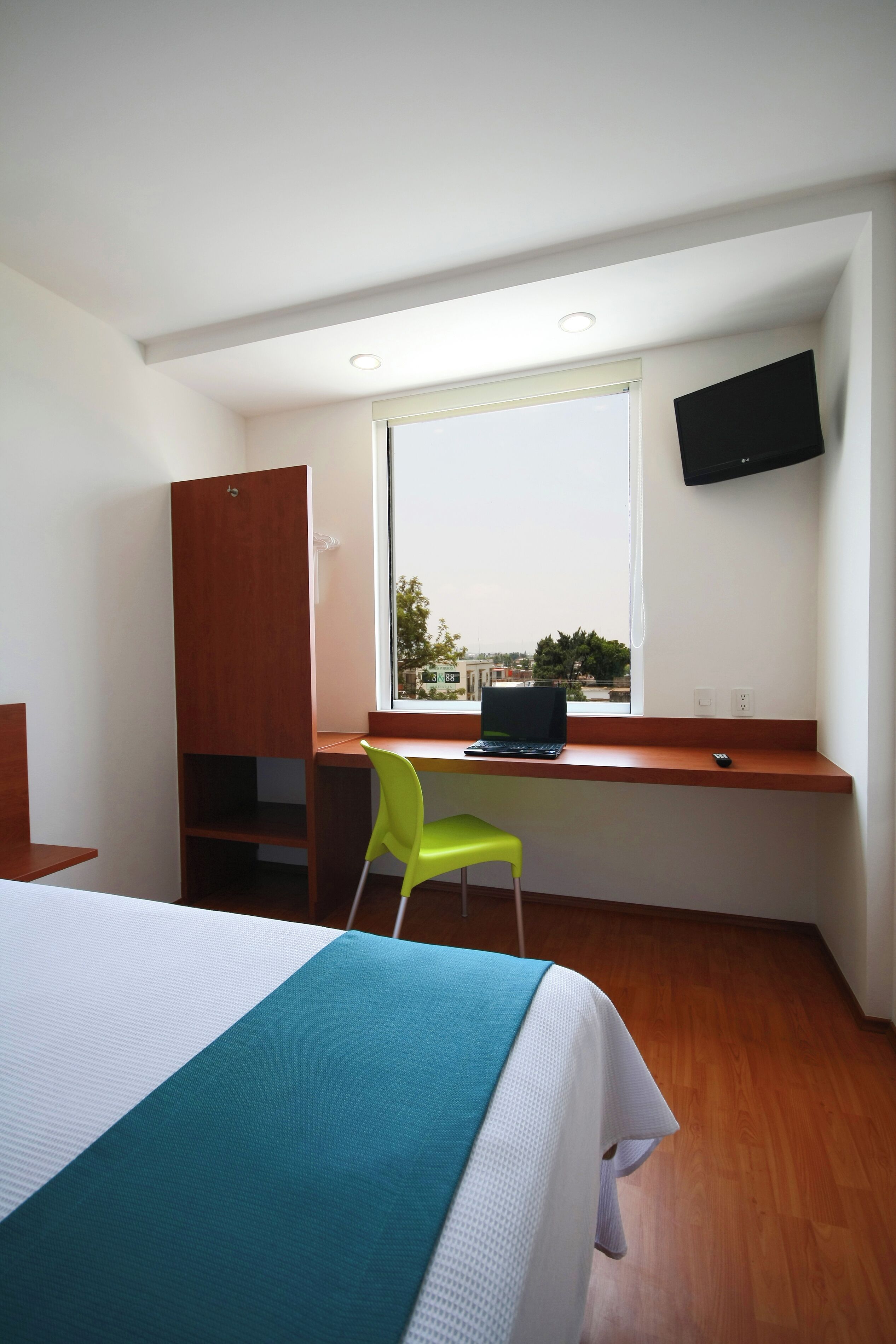 A bedroom with a bed, a desk with a laptop, a green chair, a TV mounted on the wall, and a window with a view of buildings.