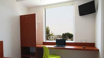 A bedroom with a bed, a desk with a laptop, a green chair, a TV mounted on the wall, and a window with a view of buildings.