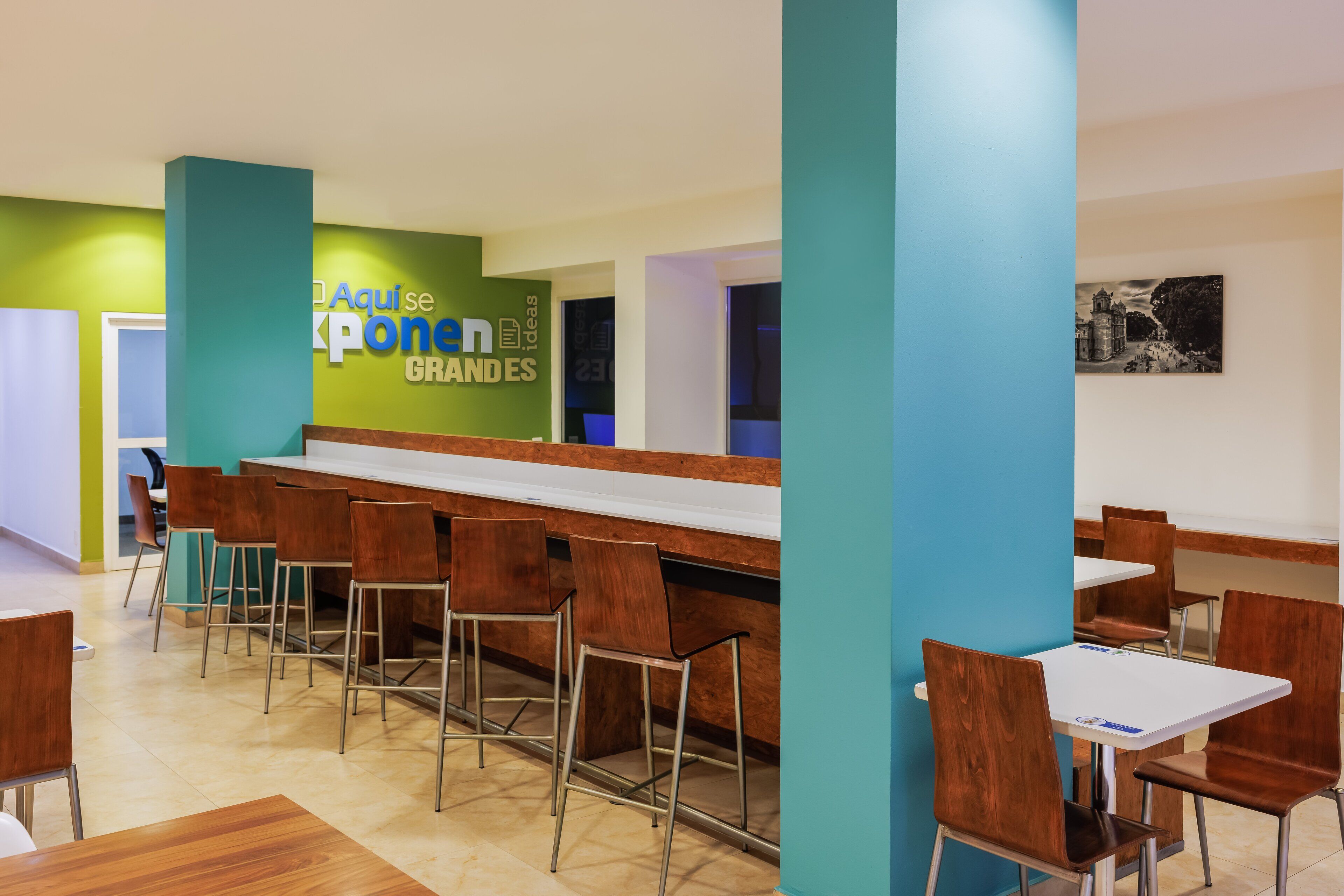 A modern cafeteria with long tables and chairs, a green accent wall, and a sign reading "Aquise Ponei Grandes".