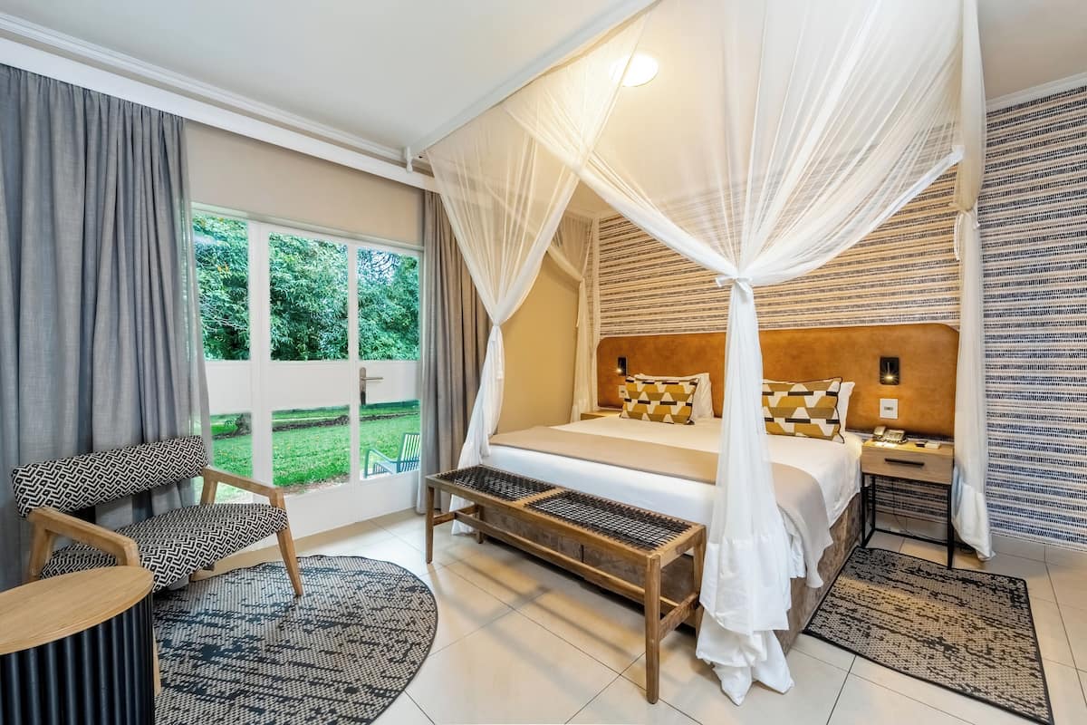 A bedroom with a bed, a chair, a bedside table, a nightstand, a window, and a view of greenery outside.